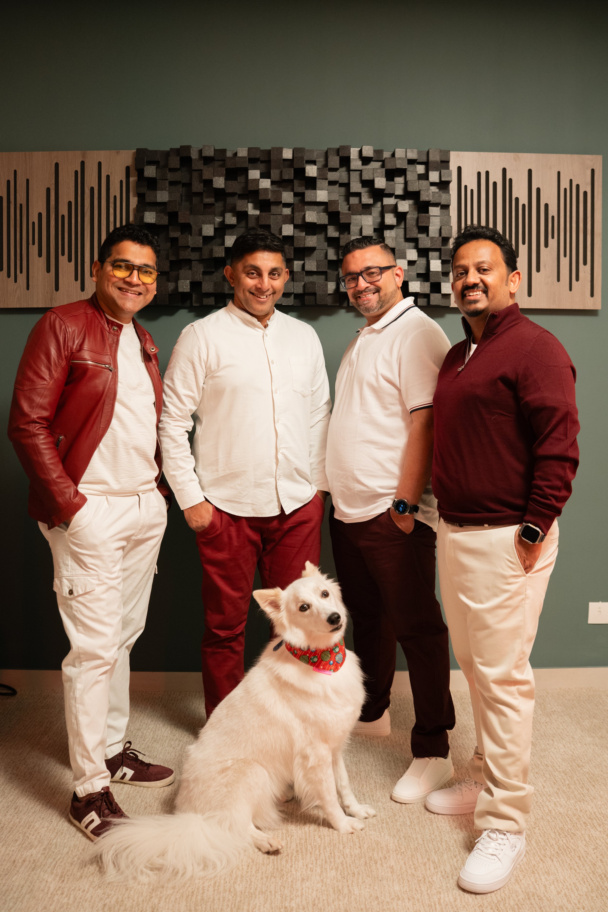 Four men posing together with a dog in front of a soundproof wall panel, smiling at the camera.