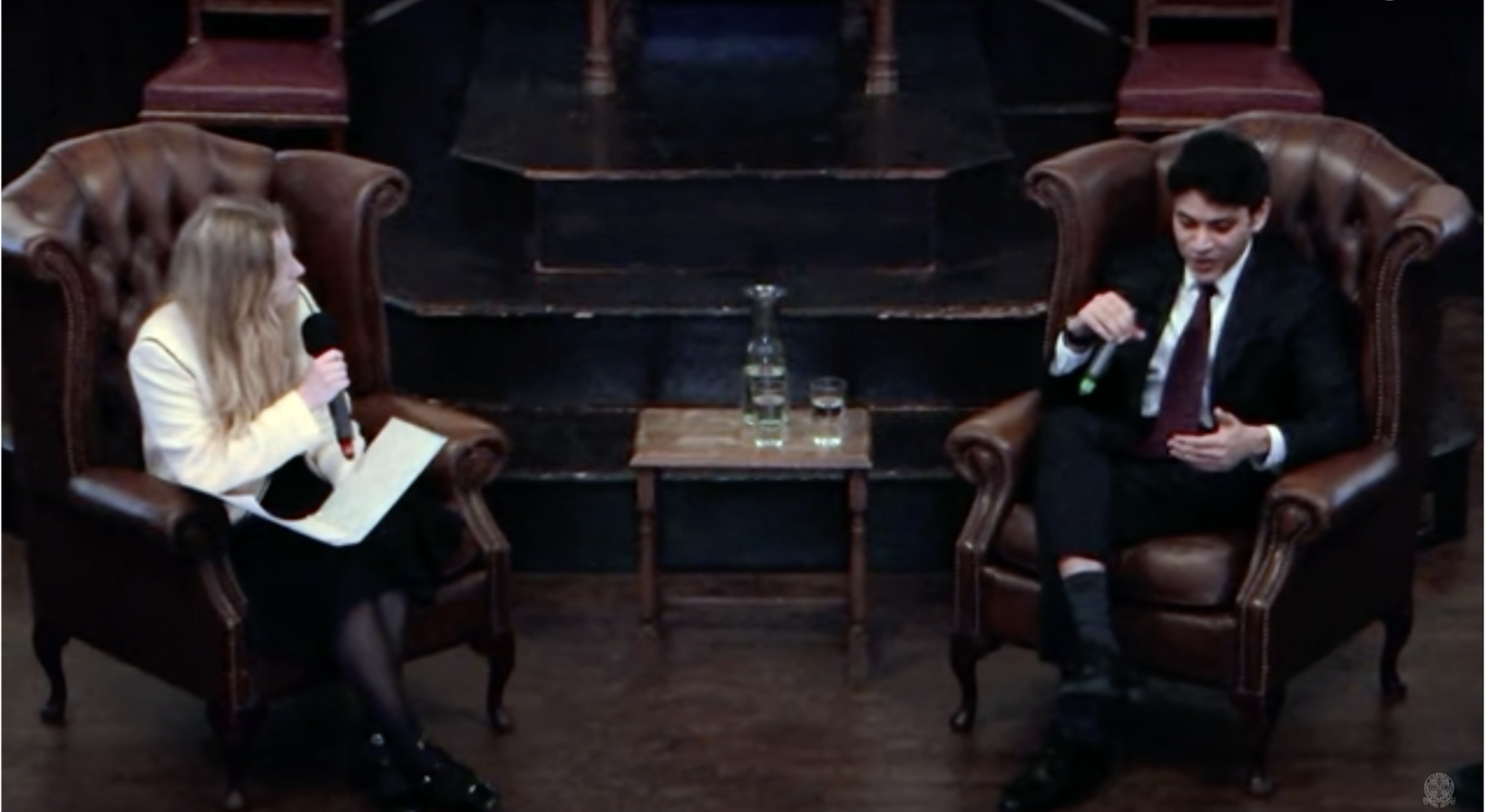 A woman and a man are sitting on separate leather armchairs facing each other in a formal setting, engaging in conversation. The woman is holding a microphone and notes, while the man is speaking. A small table between them holds a water bottle and g