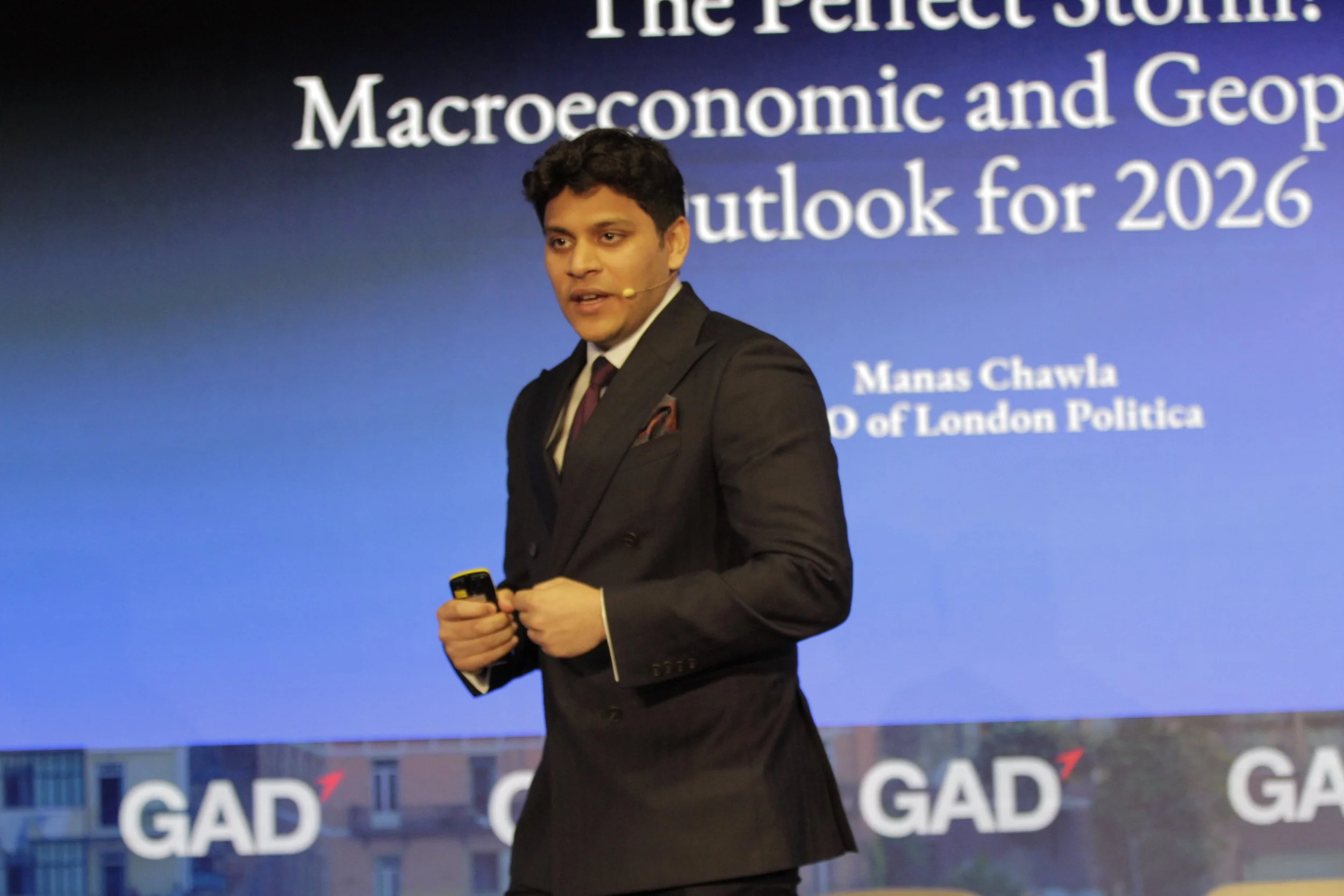 A man wearing a dark suit and a maroon tie holding a phone, standing in front of a blue background with white text about macroeconomic and geopolitical outlook for 2026.