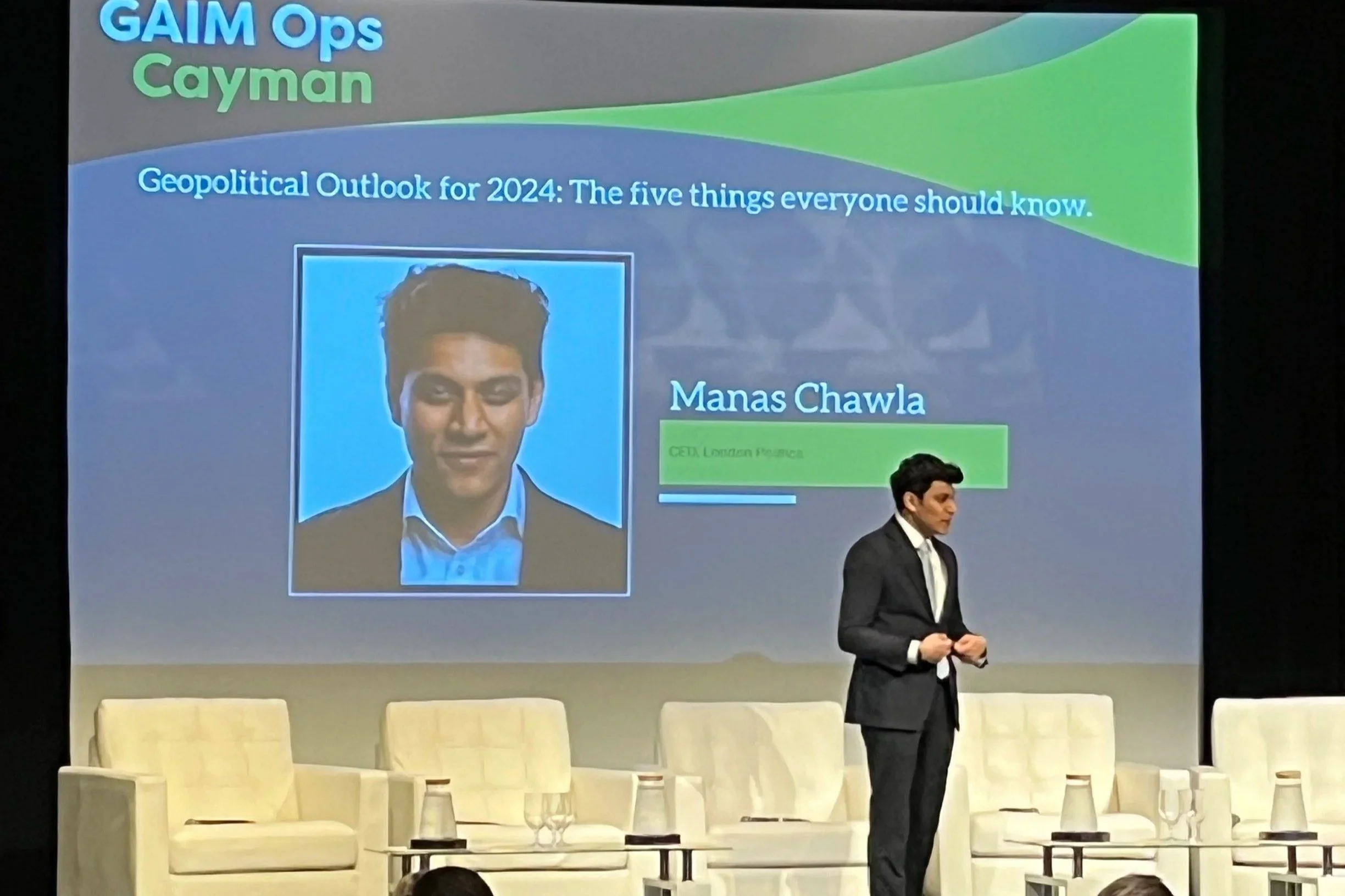 A stage presentation with a large screen displaying the title 'GAIM Ops Cayman' and the subtitle 'Geopolitical Outlook for 2024: The five things everyone should know.'