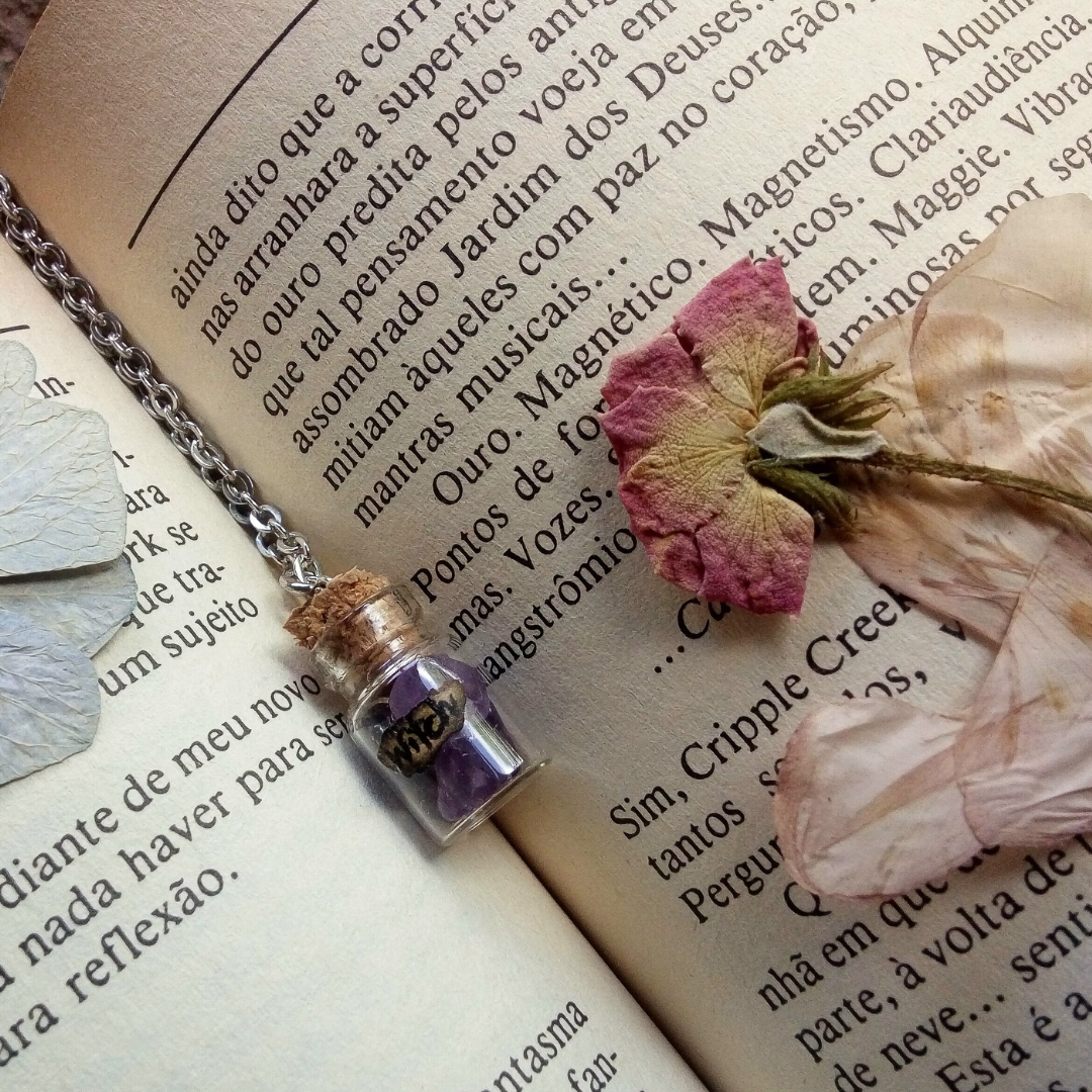 Open book with printed text, dried pink and white rose petals, small glass jar necklace with purple crystals, and a dried pink rose.