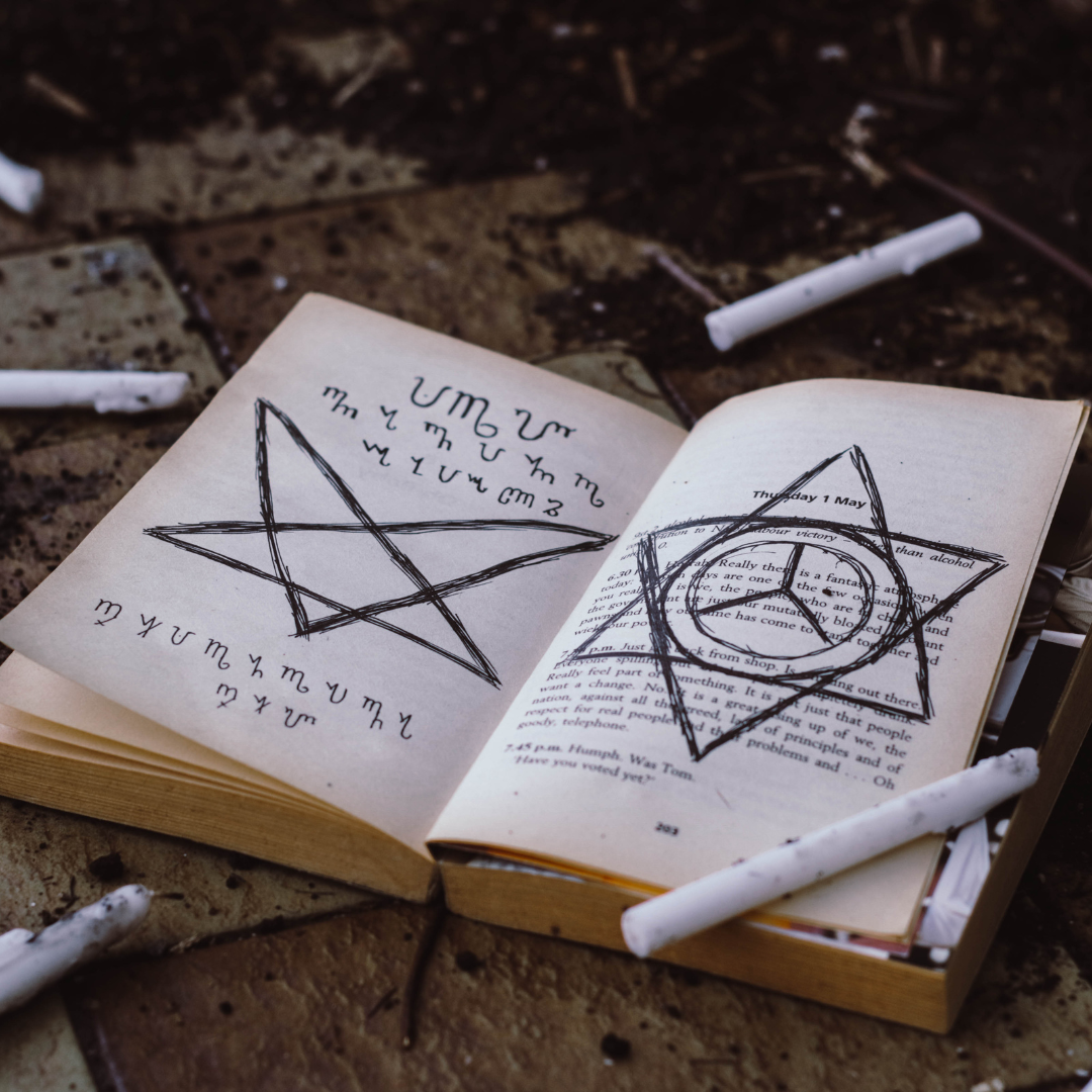 Open book on a brick surface with black pentagram drawn on pages, surrounded by used chalk sticks.