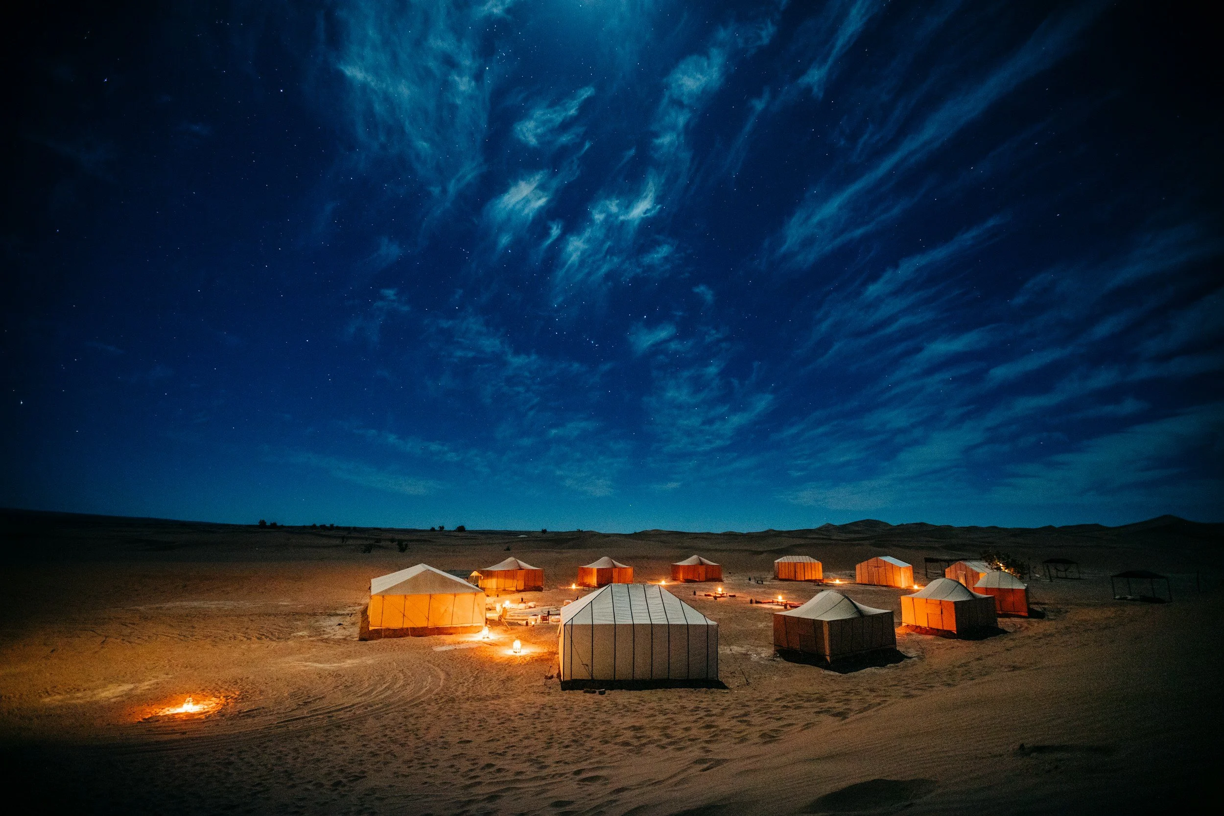 Nighttime desert scene with multiple tents illuminated by fire, under a starry sky with wispy clouds.