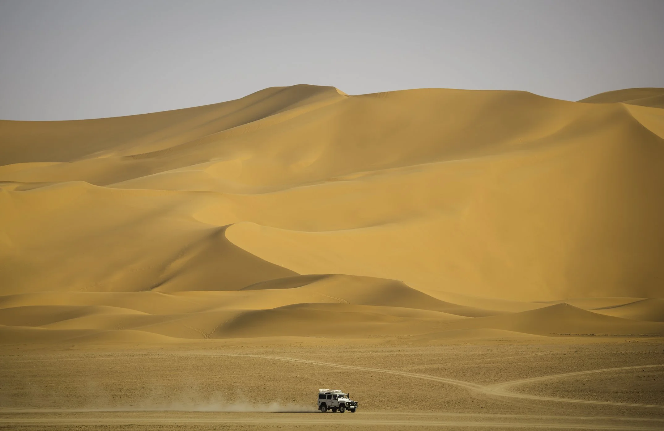 A white SUV driving across a desert landscape with large sand dunes in the background.
