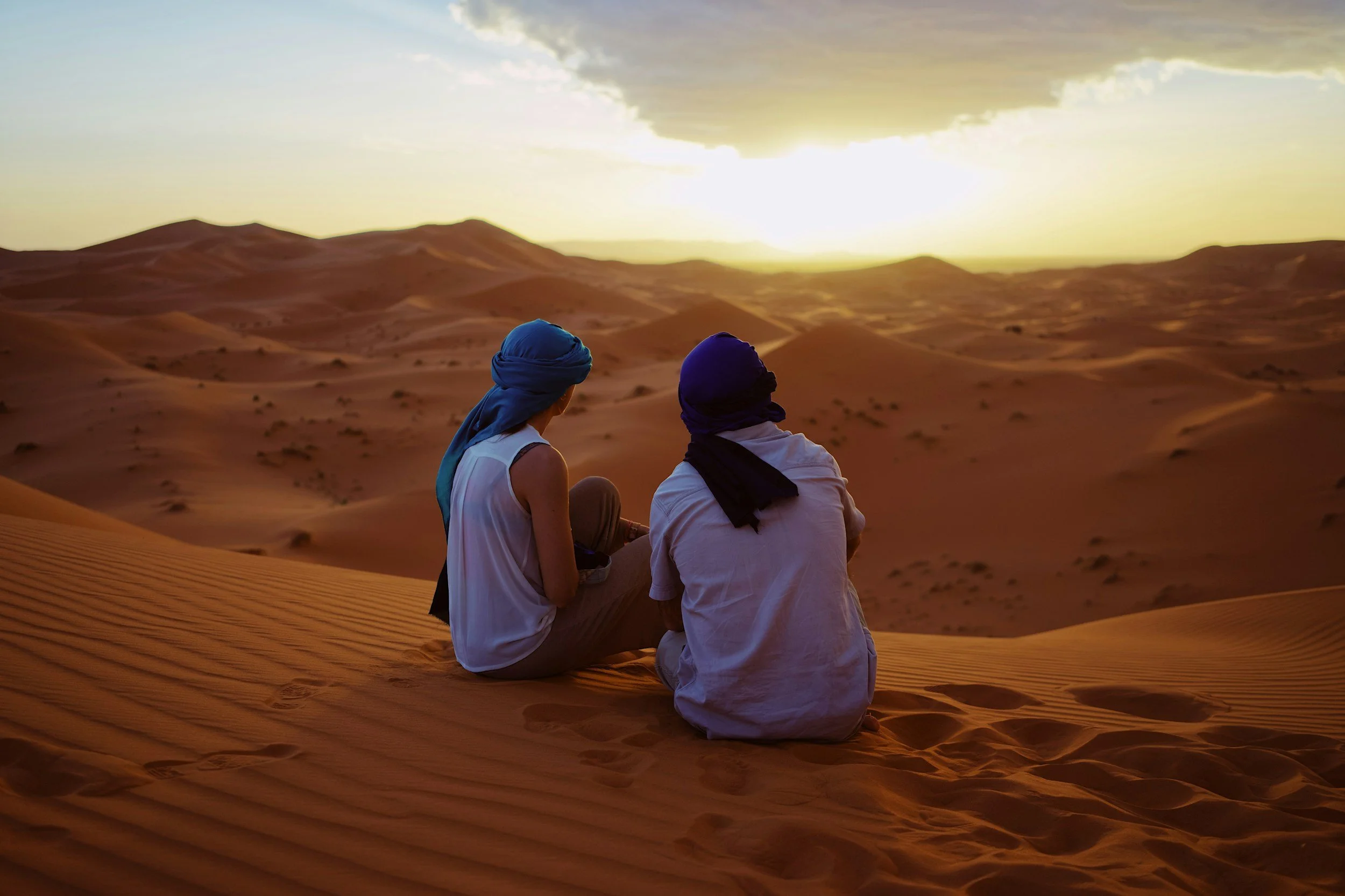 Two people with head coverings sitting on a sand dune in a desert landscape at sunset, looking towards the horizon.