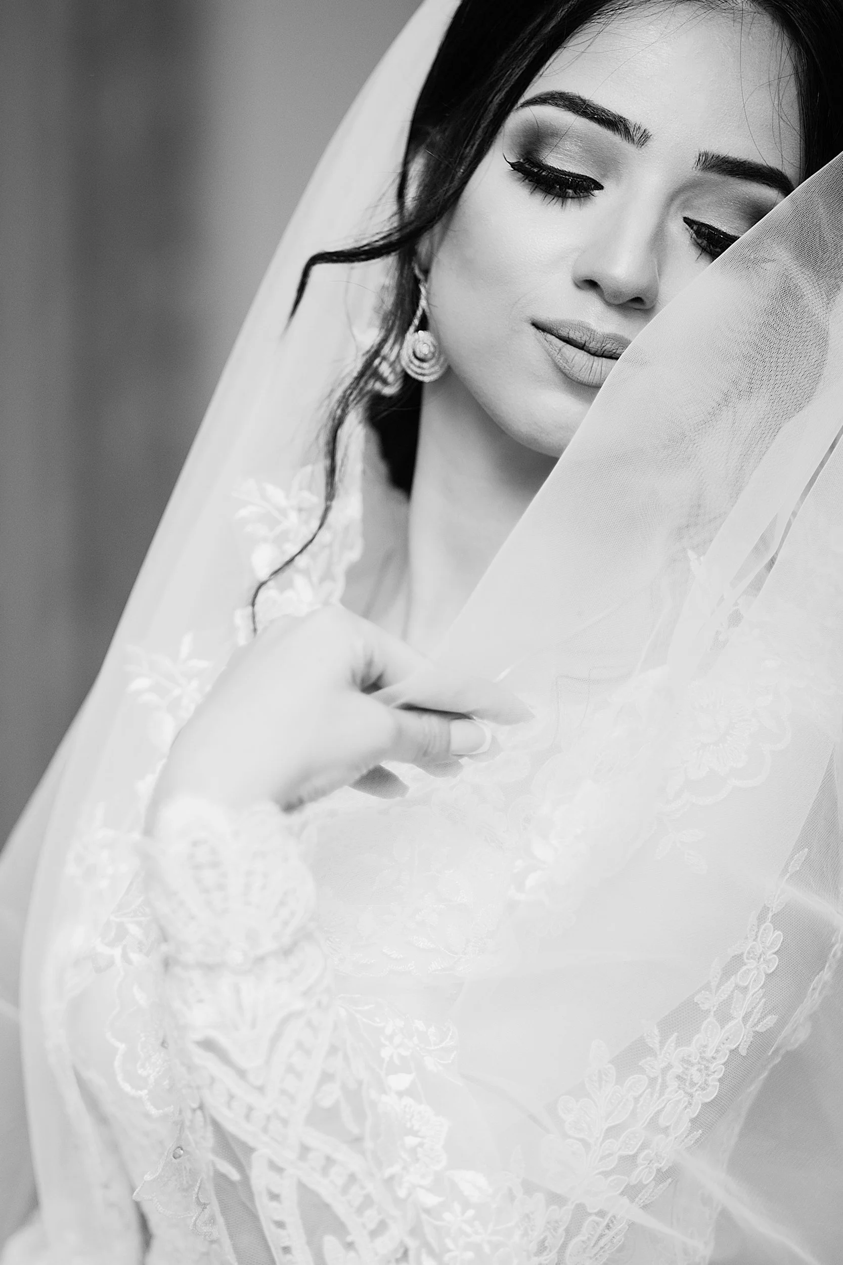 A close-up black-and-white photo of a woman with closed eyes, makeup, and earrings, holding a lace-trimmed veil or fabric.