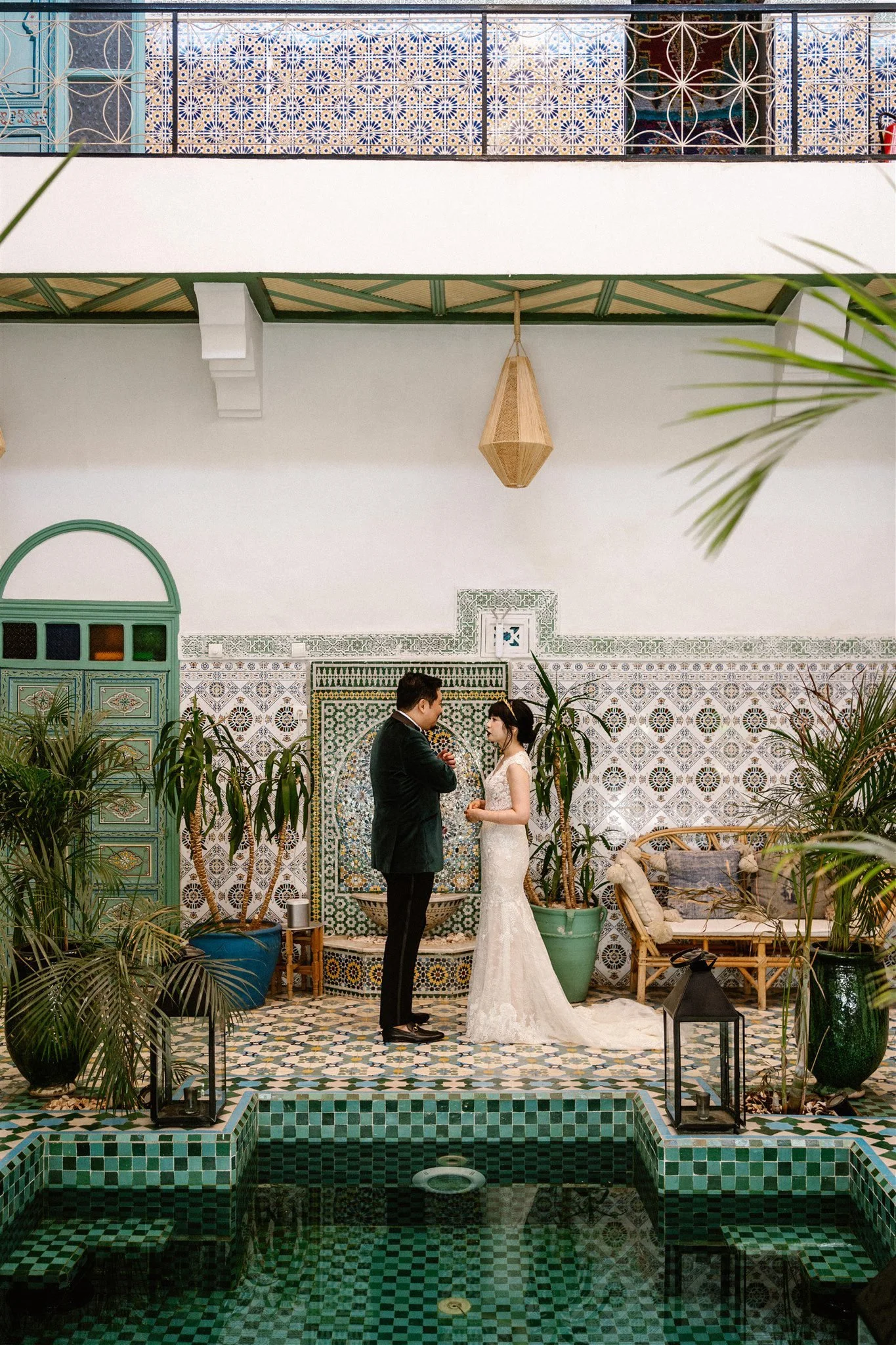 A bride and groom exchange vows in a Moroccan-inspired indoor setting with intricate tilework, lush plants, and a reflective green-tiled pool in the foreground.