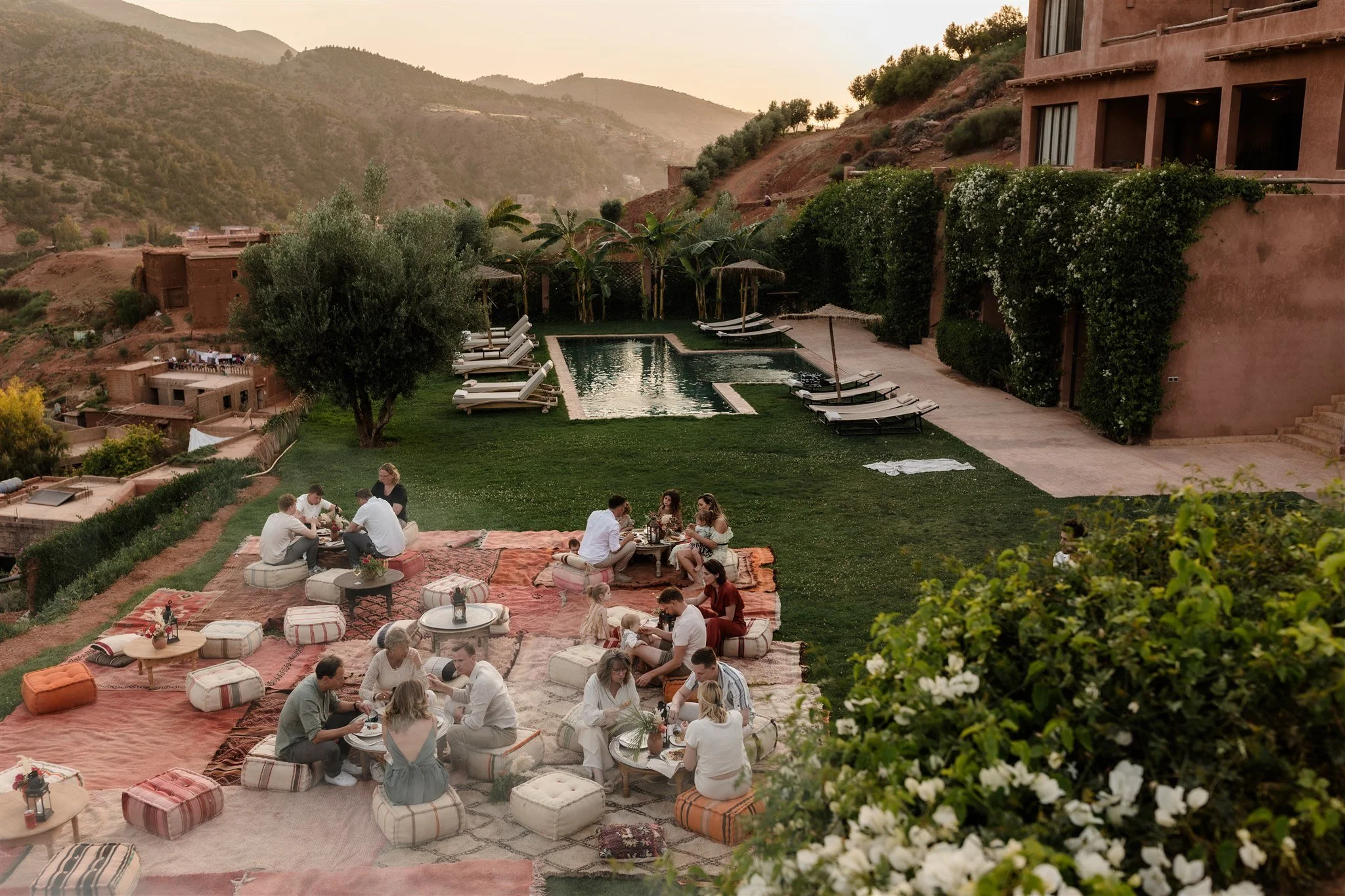 People gathered outdoors at a hillside of the Atlas mountain gathering, sitting on cushioned rugs, with a swimming pool in the background, surrounded by mountains and greenery during sunset.