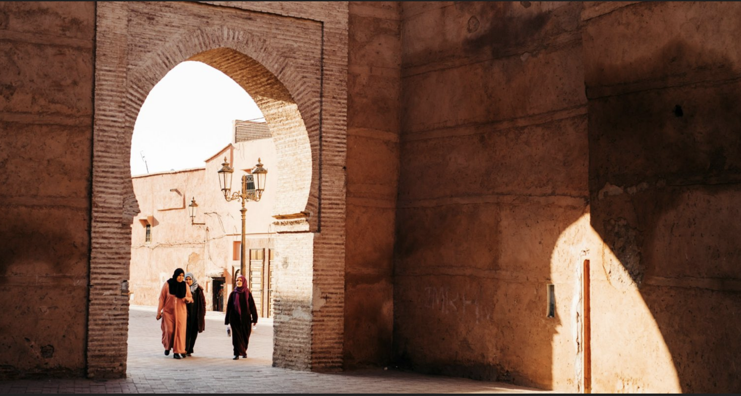 People walking through the ancient city archway of Fes Morocco made of brick with traditional street lamps and old buildings in the background.