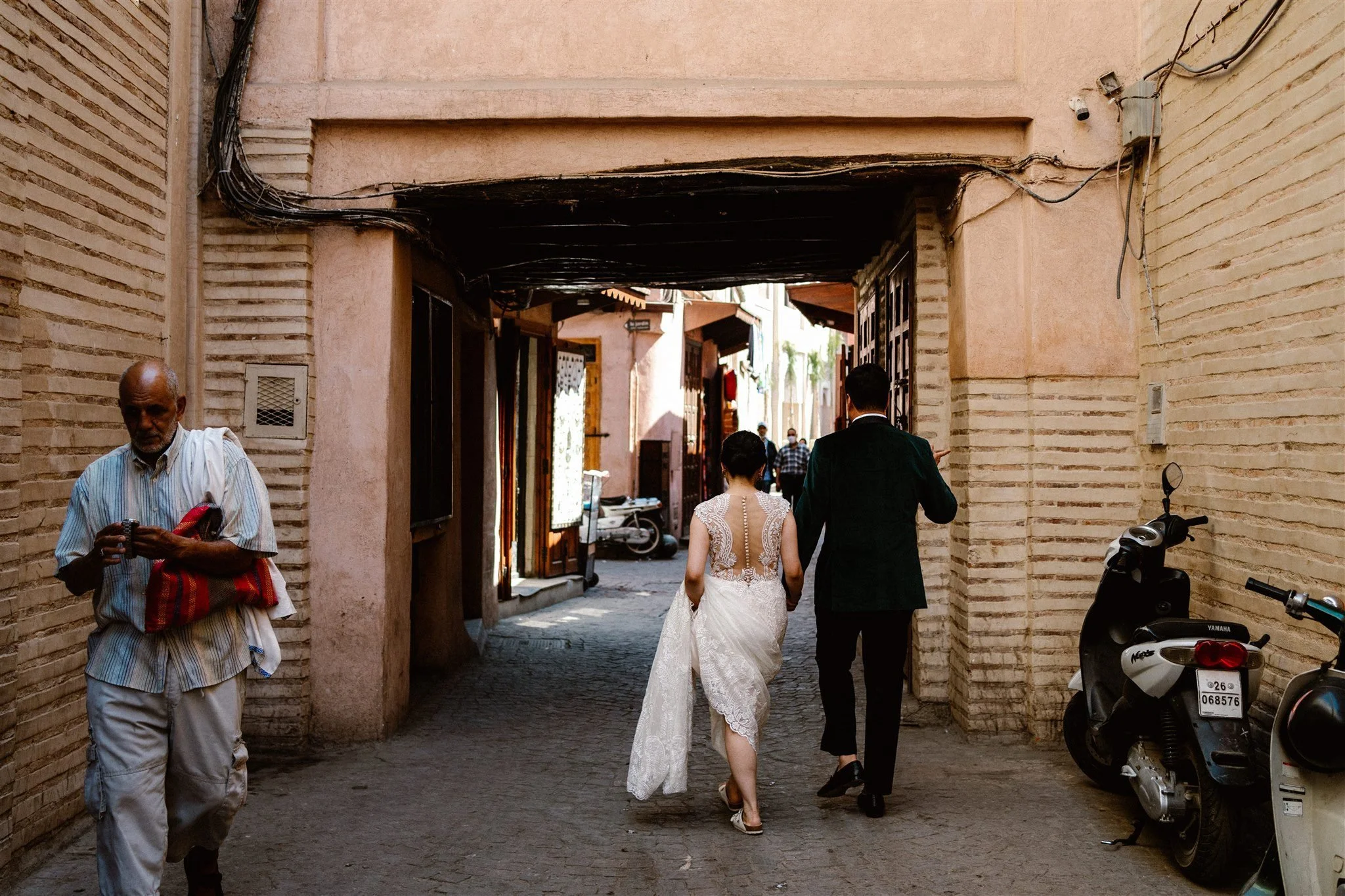 A wedding couple walking through a narrow alleyway with brick walls on either side. The bride is wearing a white dress and the groom is in a dark suit. There are other people and parked motorbikes in the background, and an older man holding a red bag is walking on the left side of the frame.