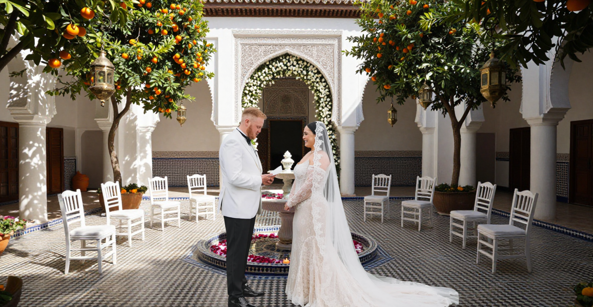 A couple in wedding attire standing in front of a decorative archway, exchanging vows or reading vows, in an outdoor courtyard decorated with white chairs, orange trees, and floral arrangements.
