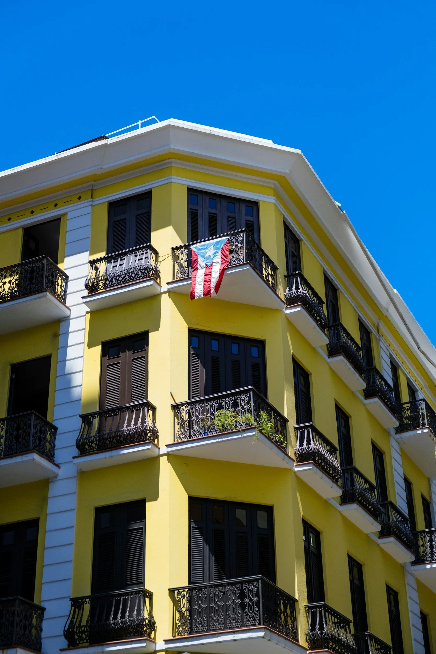 A yellow apartment building with black balconies and a Puerto Rican flag hanging from one of the balconies under a clear blue sky.
