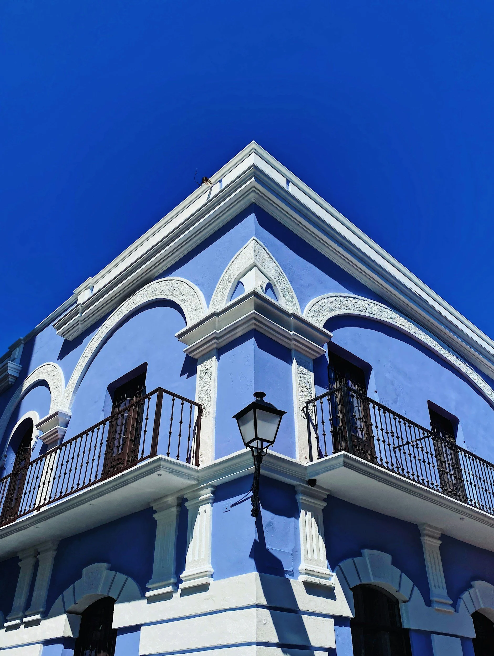 Blue building with white accents, a corner balcony with black wrought iron railings, and a street lamp casting a shadow, against a clear blue sky.