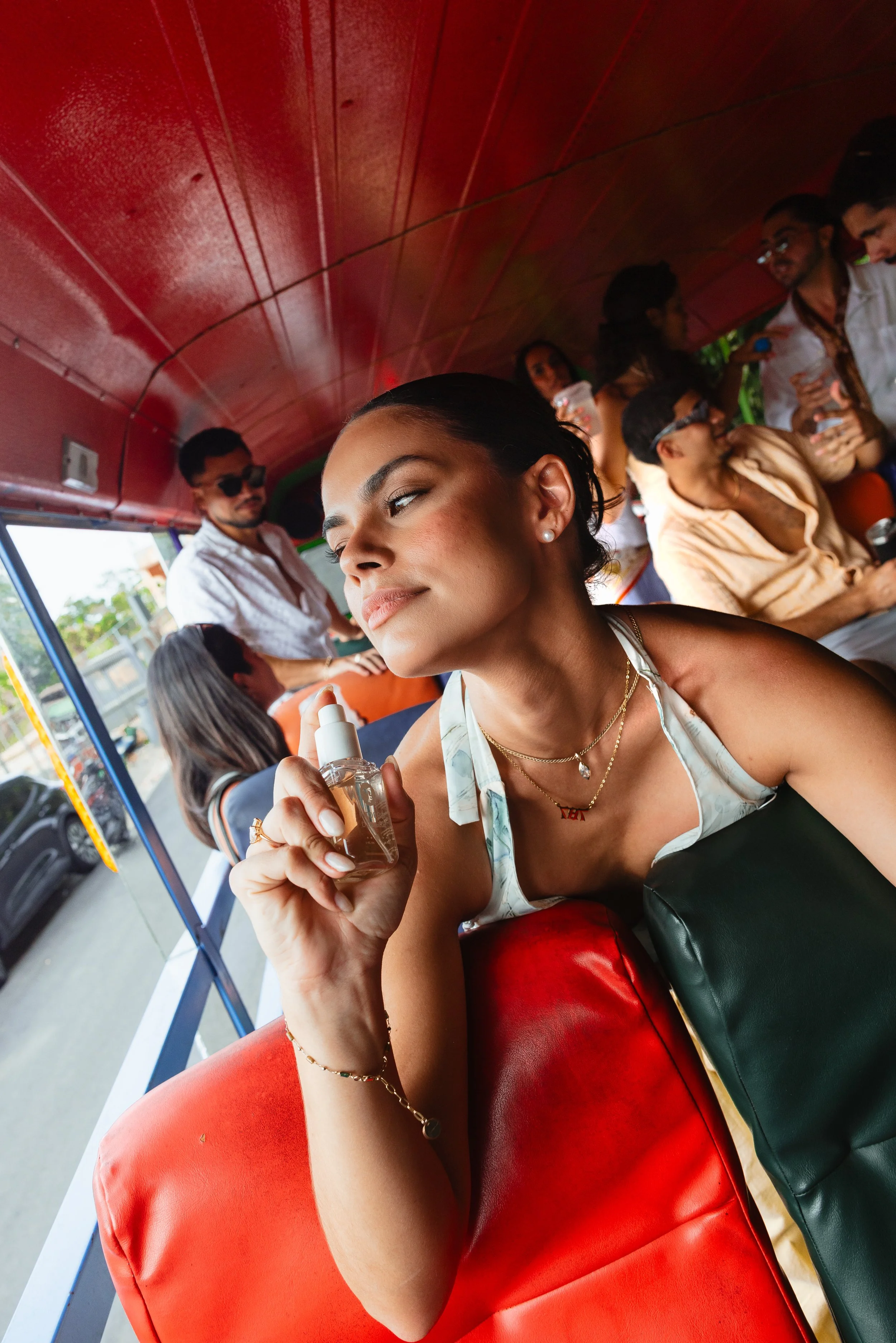 A woman with dark hair, wearing pearl earrings and jewelry, holding a spray bottle, sitting on a red seat on a bus or train with other passengers, some standing and holding drinks, under a red curved ceiling.