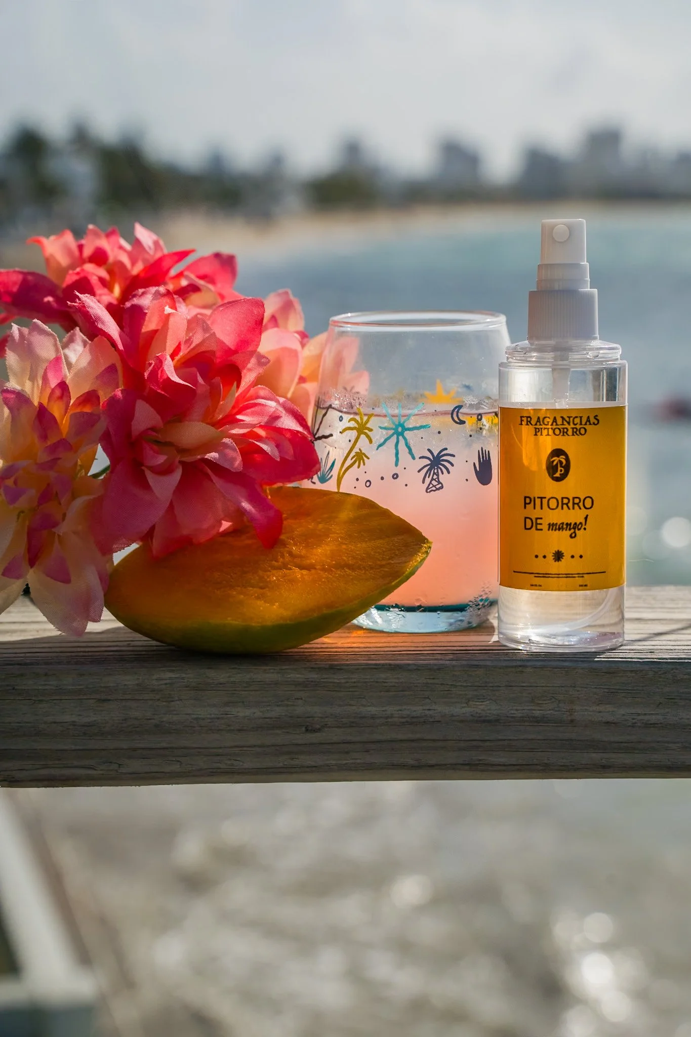 A tropical scene with pink flowers, a sliced mango, a glass with a decorative drink, and a spray bottle of mango mist on a wooden surface near the beach.