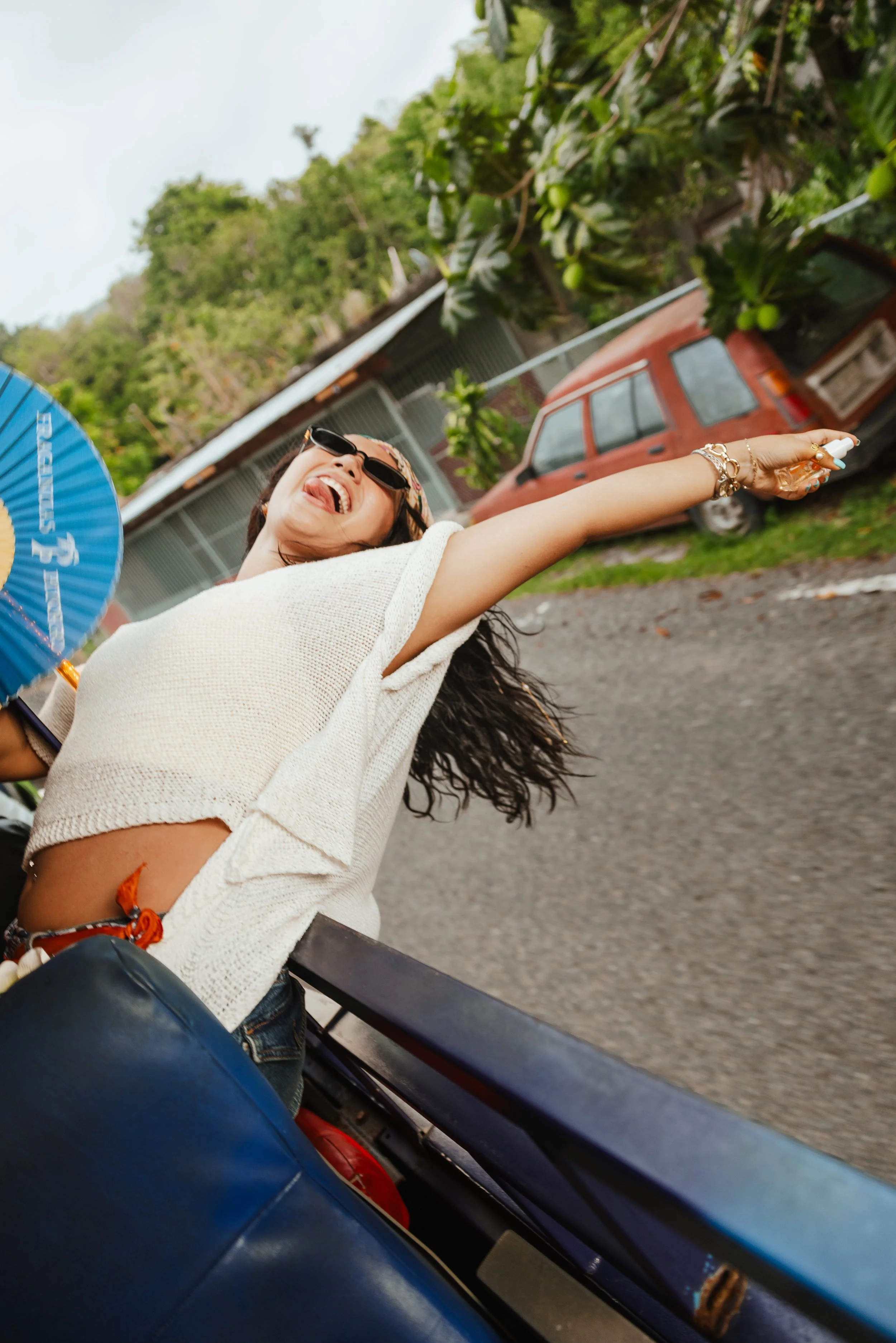 Woman with long hair wearing sunglasses and a white top, sitting outdoors on a bus or similar vehicle, smiling and leaning back with her tongue out and her arm extended, holding a small spray bottle. There is a background of green trees, cars, and a building.