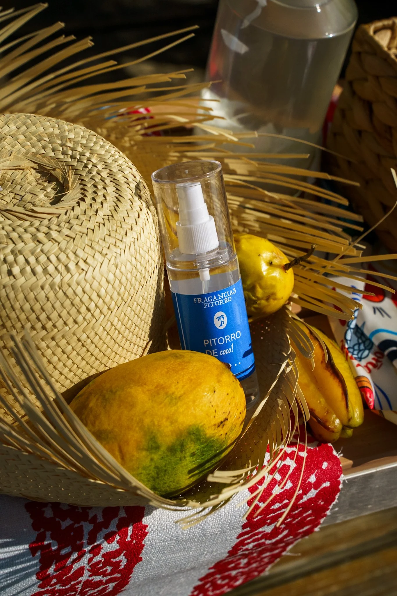 A woven straw hat, a small spray bottle labeled 'PITORRO de coco!', two mangoes, a banana, a glass of water, a cloth with a red and white pattern, and some palm fronds on a table.