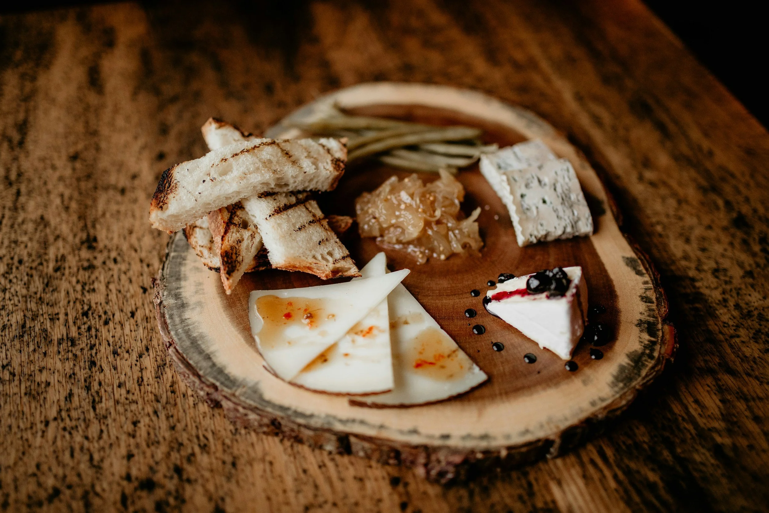 A wooden platter serving three different types of cheeses, a thin cut hard cheese, with some chilli sauce, a wedge of soft cheese with berry compote and a blue cheese. served with some caramelised onions and charred slices of artisan bread