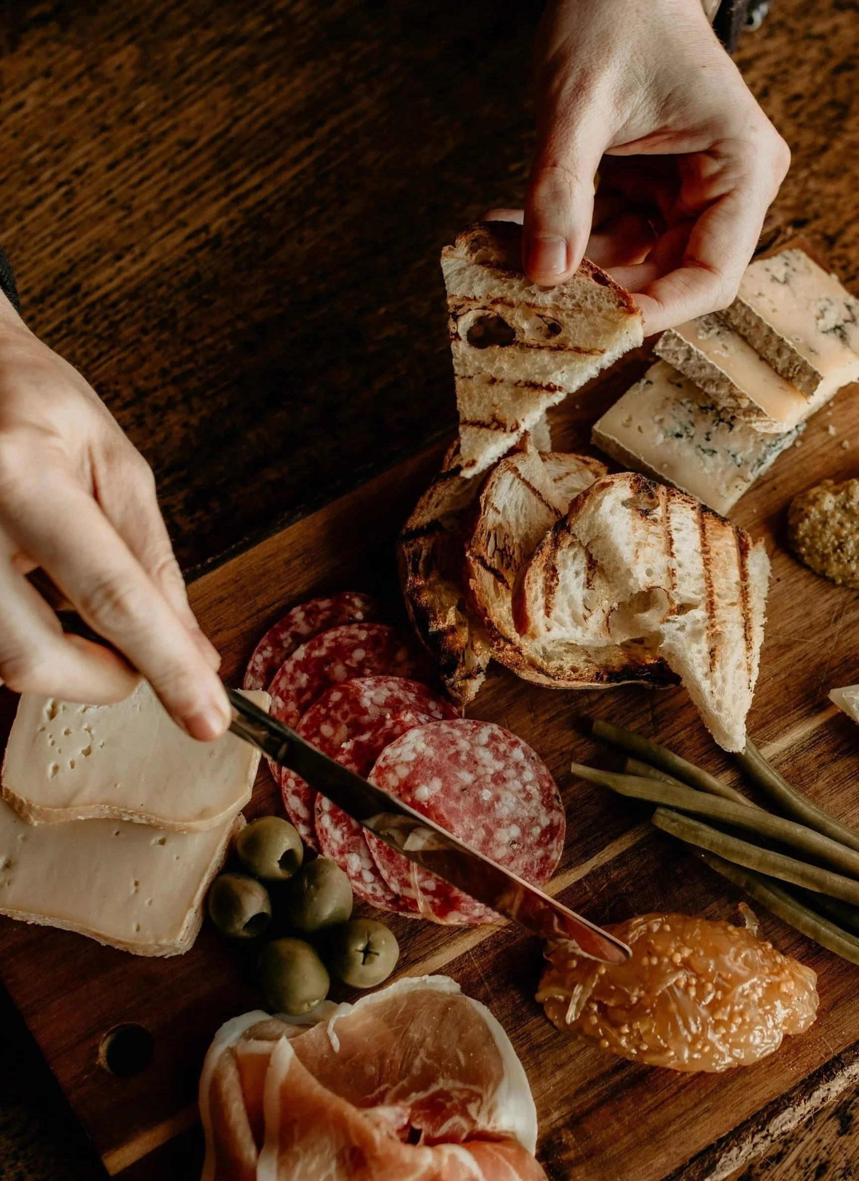 A charcuterie board with slices of bread, cheese, cured meats, green olives, a dollop of mustard, and some green beans, on a wooden surface.
