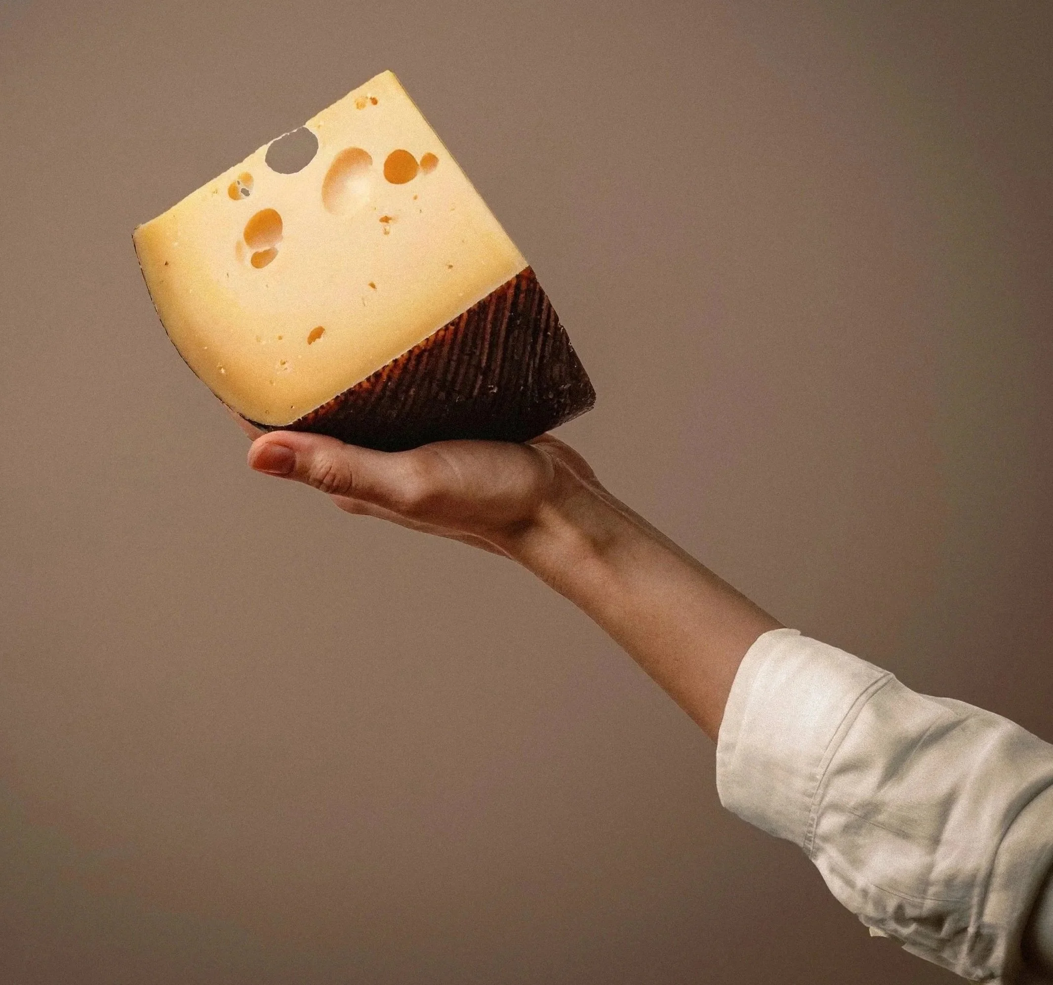 Hand holding a large wedge of Swiss cheese with holes against a neutral background.