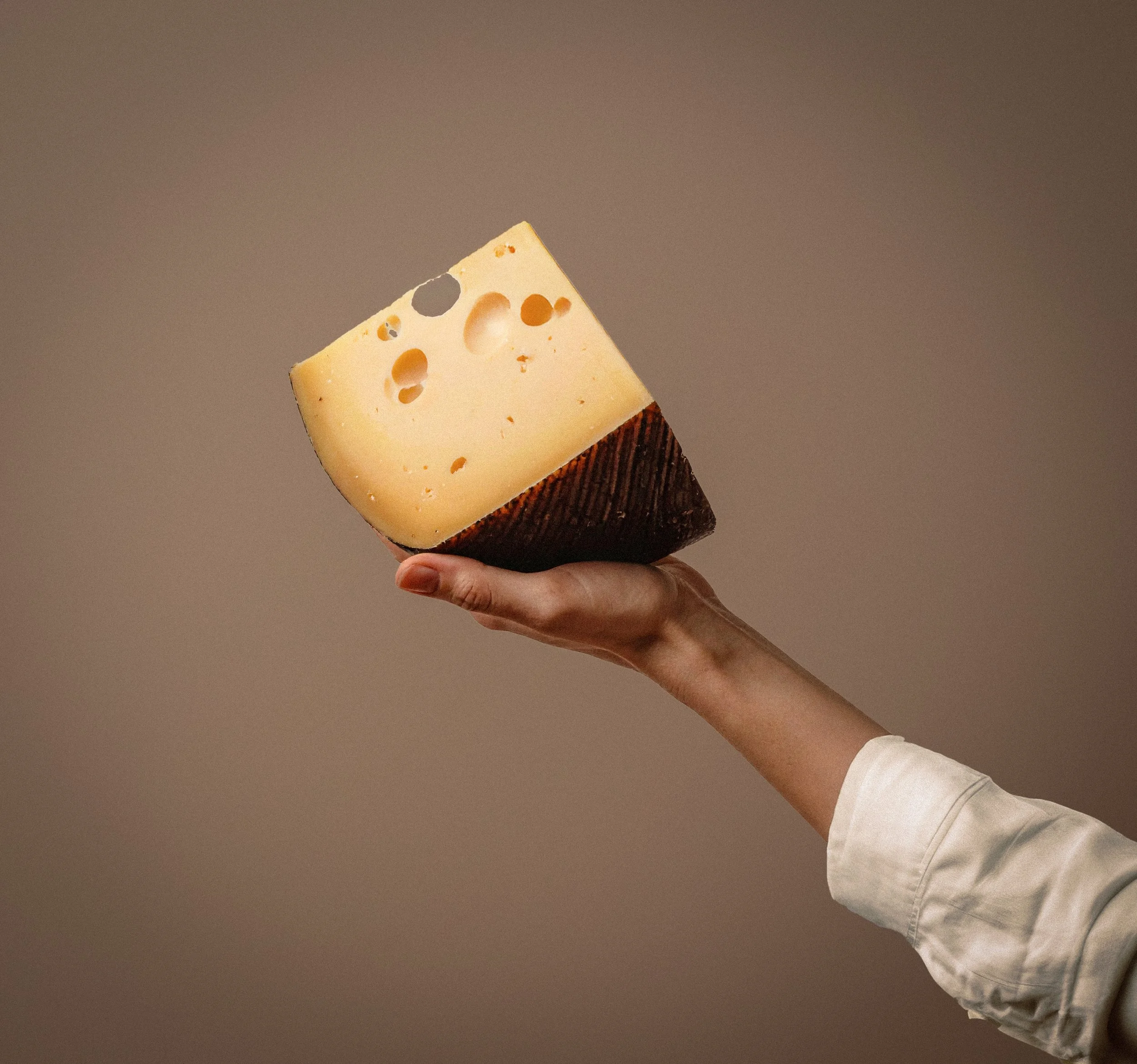 Hand holding a wedge of Swiss cheese with a brown rind, against a plain background.