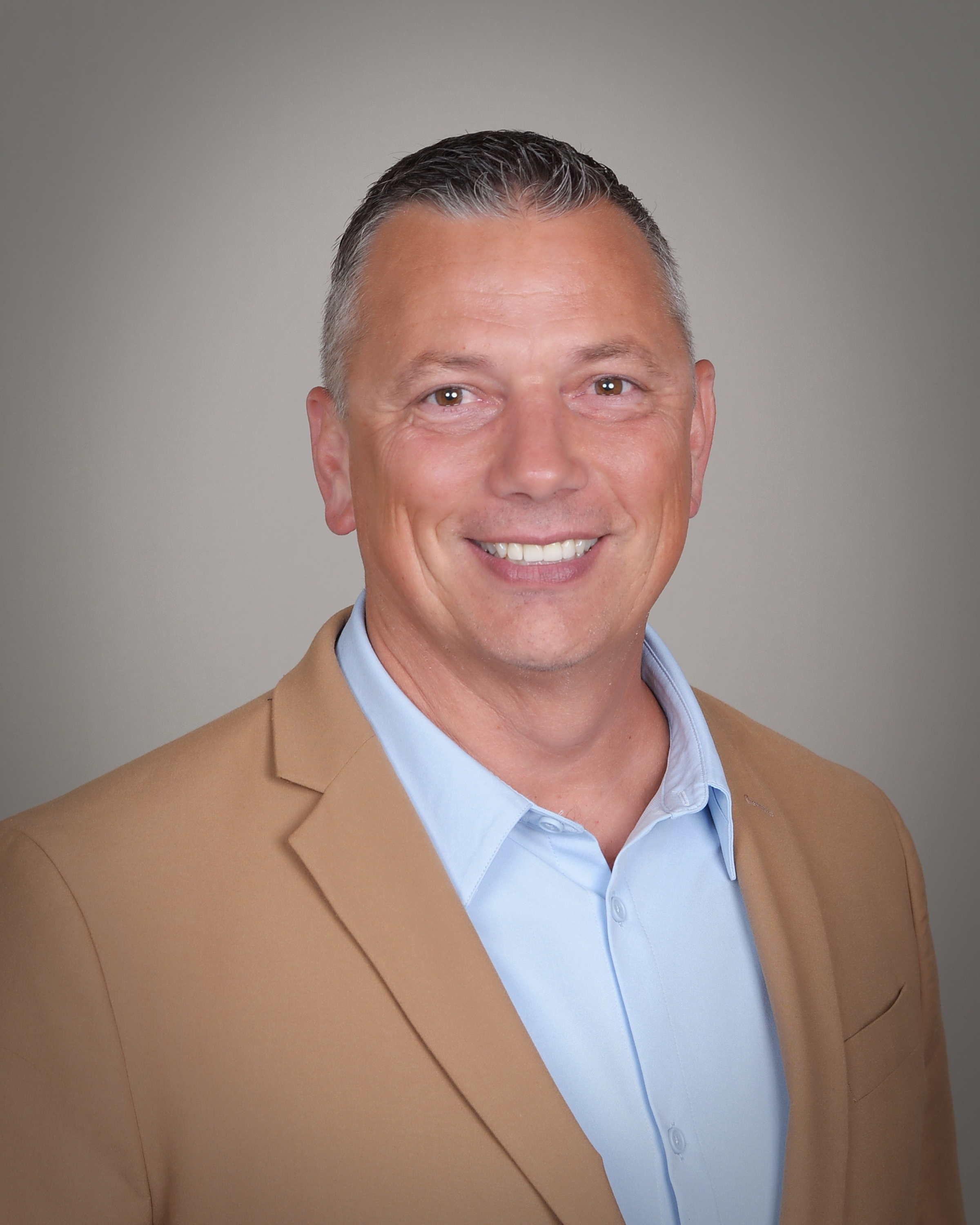 Close-up of a smiling man with short gray hair, wearing a tan blazer over a light blue collared shirt, against a neutral gray background.