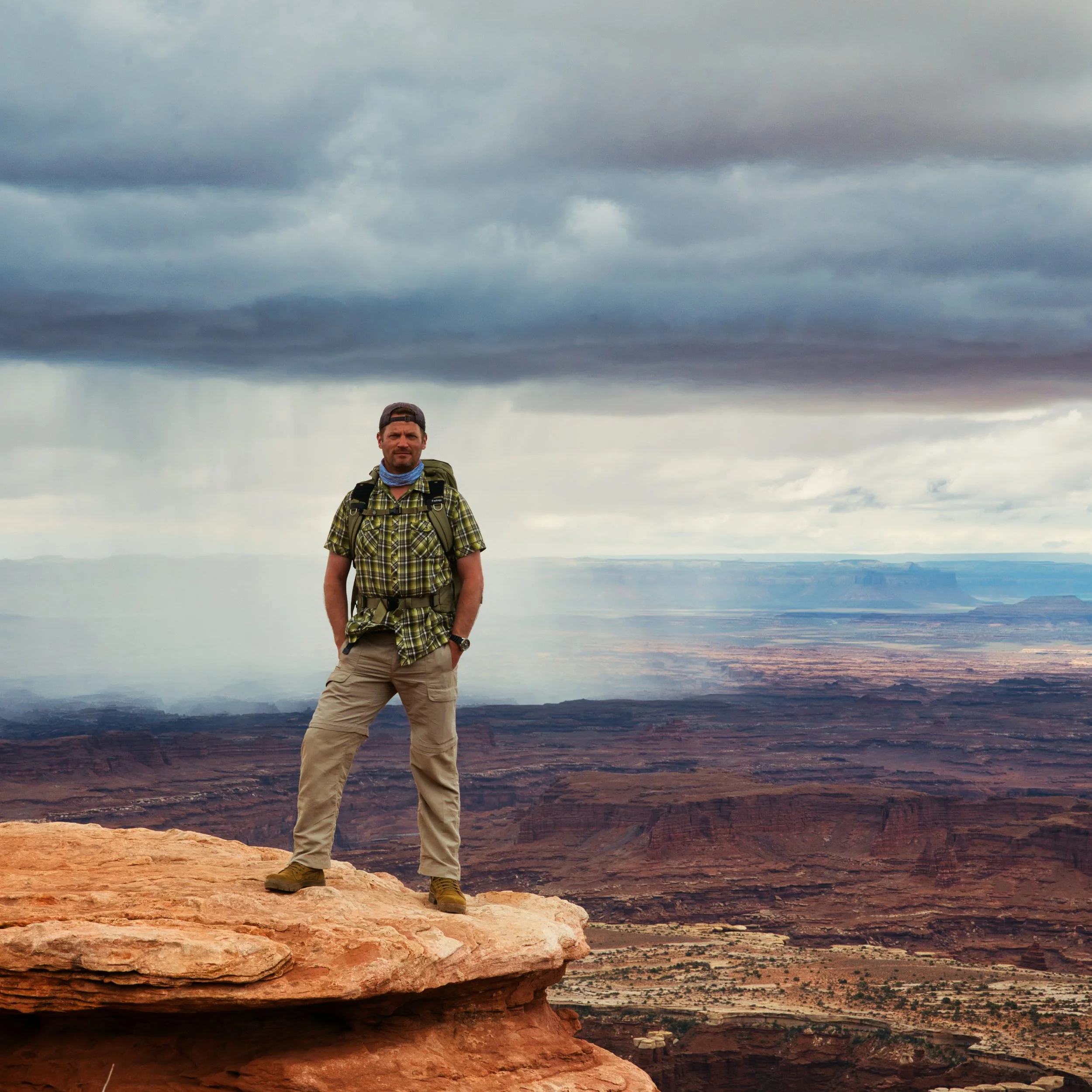 A man standing on a rock ledge overlooking a vast canyon landscape under a cloudy sky.