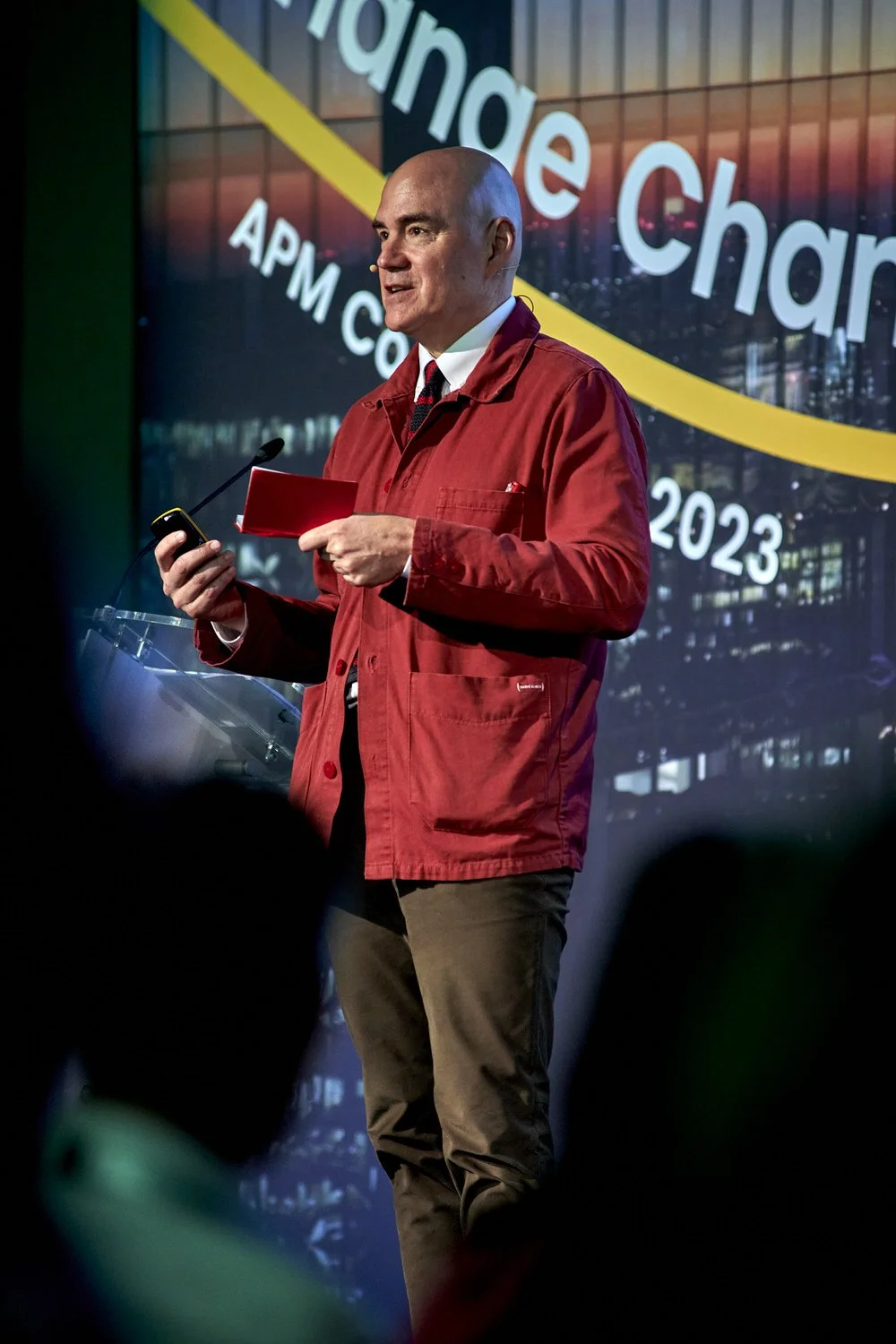 Man in red jacket speaking on stage at an APM event.