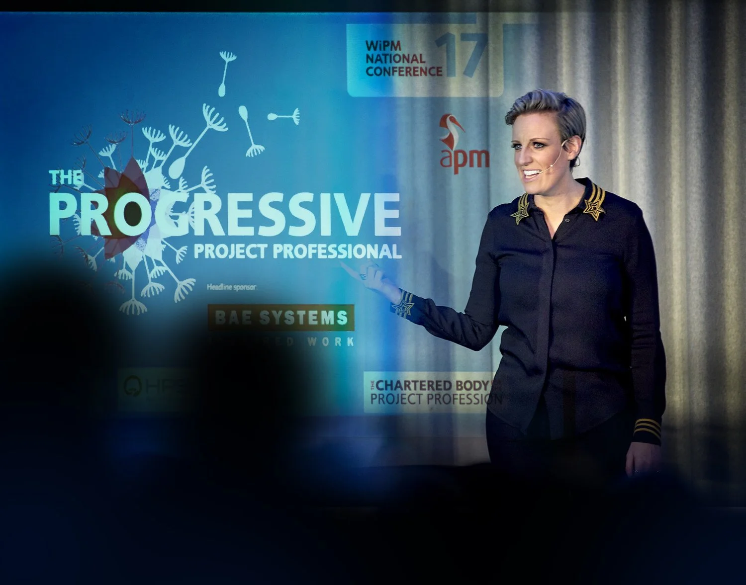 Presenter delivering a talk at a progressive business event, audience blurred in foreground.