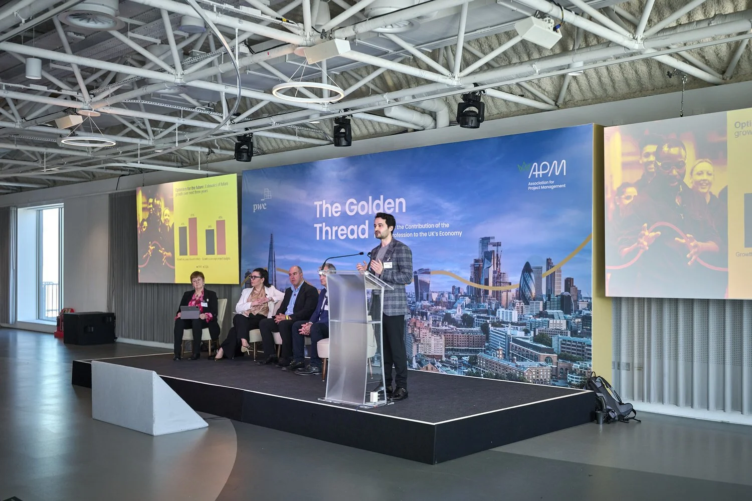 Male speaker standing on stage with a panel of four seated behind him.