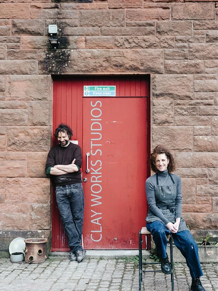Two artists smiling outside a red door with the words 'Clay Works Studios' written on it, set in a brick wall. One man is leaning against the wall with arms crossed, and one woman is sitting on a stool wearing an apron.