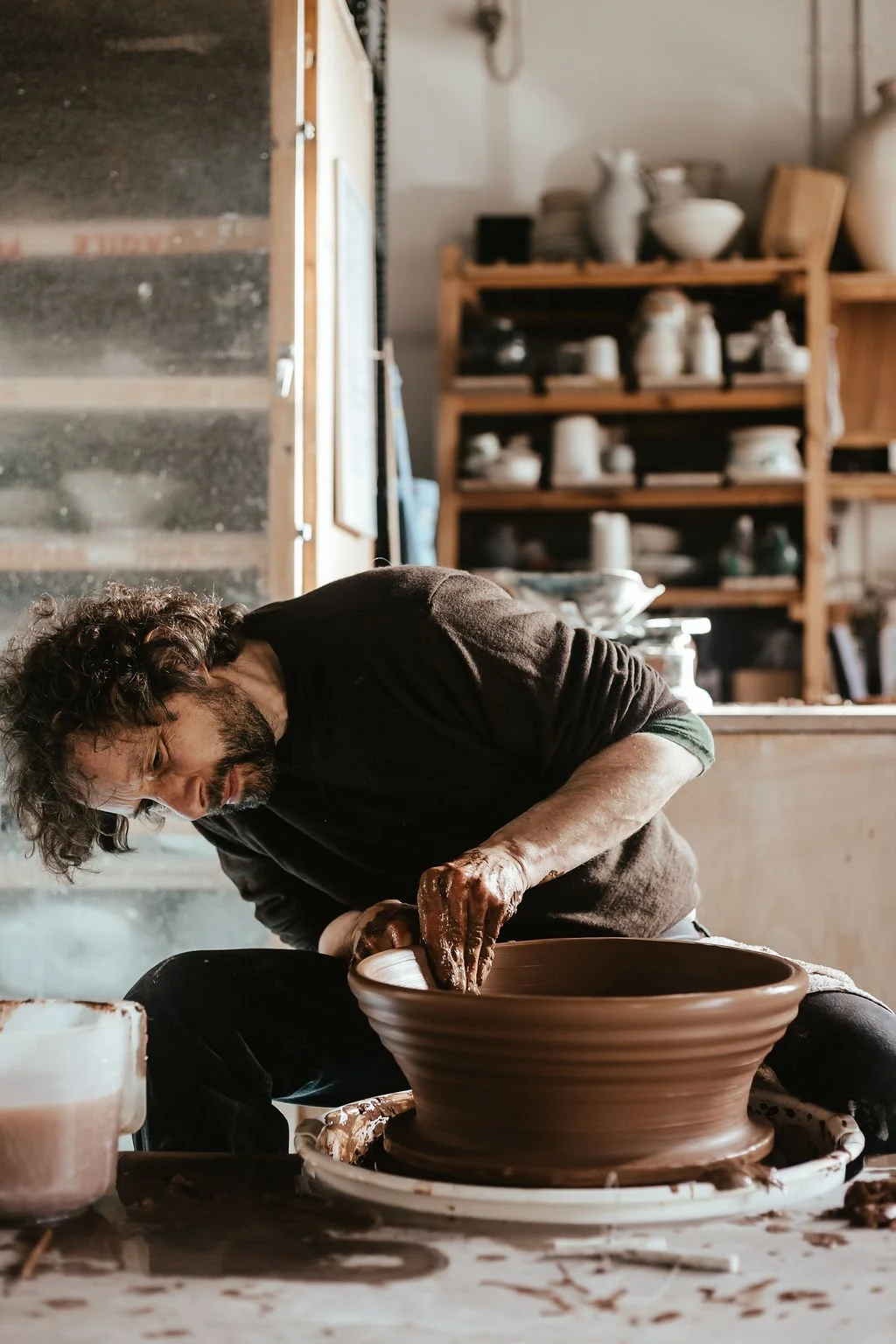 A man shaping a large clay pot on a pottery wheel inside a studio with shelves of pottery in the background.