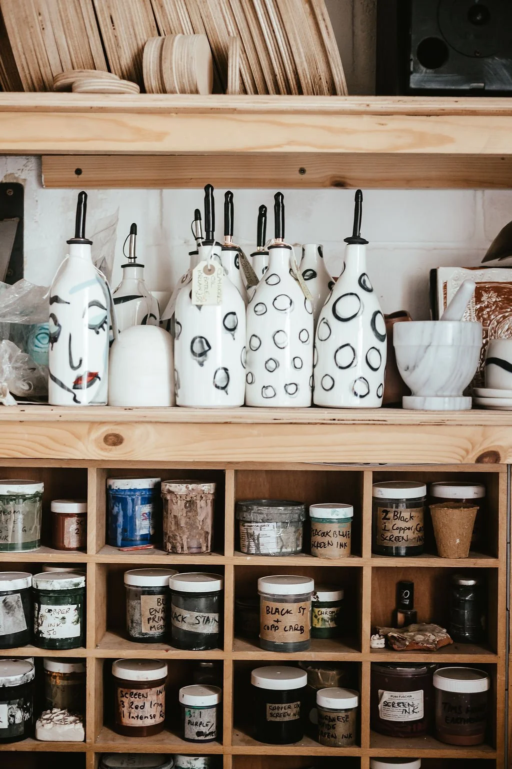 Shelf with decorative bottles featuring abstract face and circle designs, alongside bowls and ceramic items, with paint jars below.