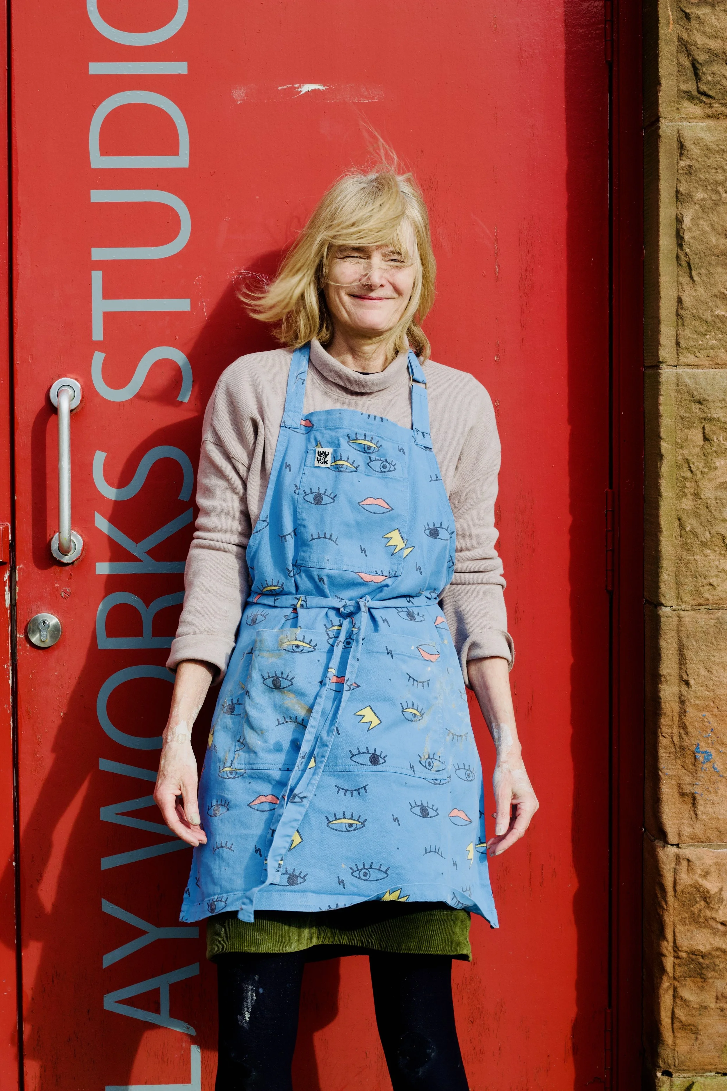 A woman with blonde hair standing in front of a red wall, wearing a blue apron with eye and lightning bolt patterns, smiling gently.