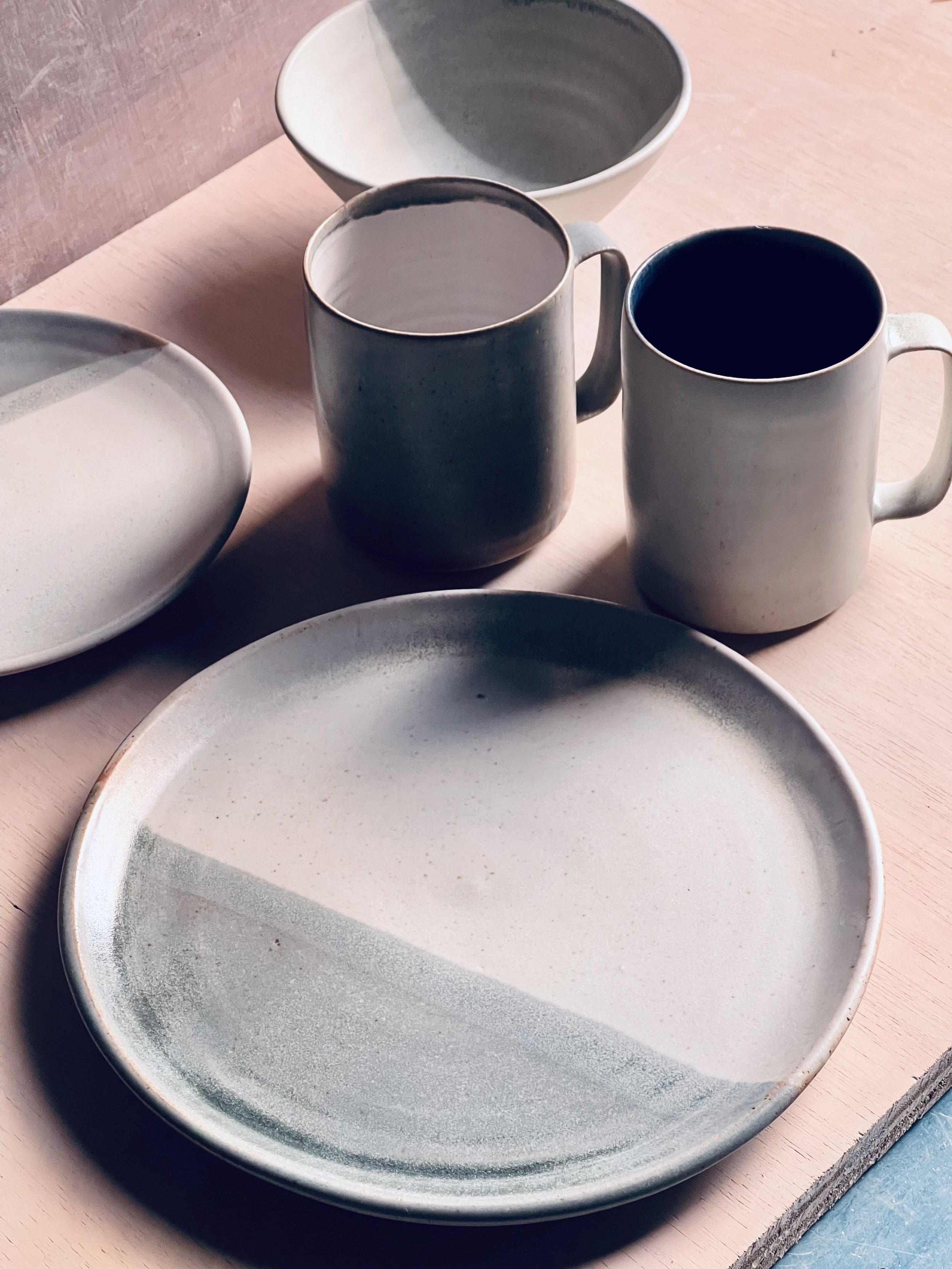 Set of ceramic plates, mugs, and bowls in neutral tones on a light wooden table.