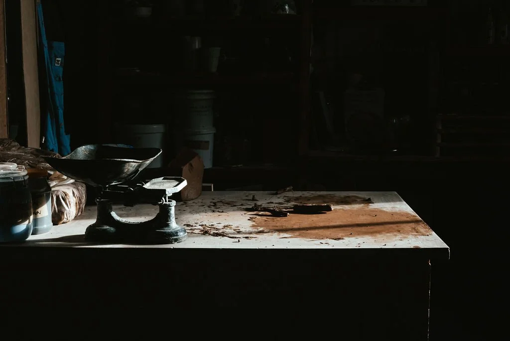 A dusty and cluttered workbench or table in a dark workshop, with a vintage scale, jars, and other objects.