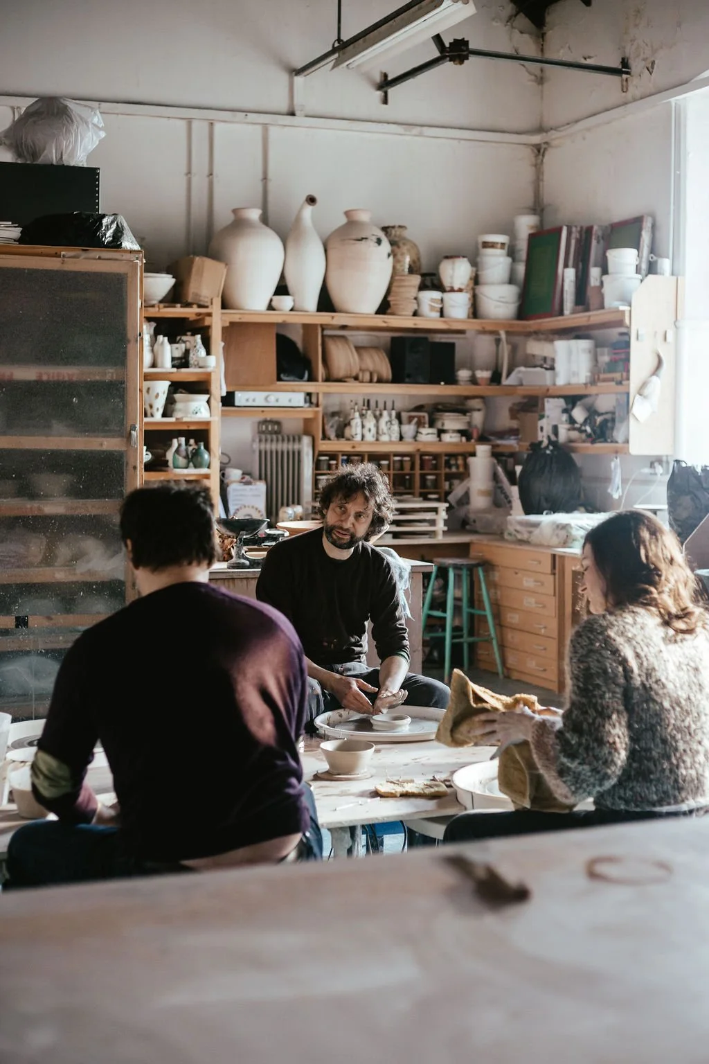 Three people seated around a table in a ceramics studio, with shelves of pottery and tools in the background.