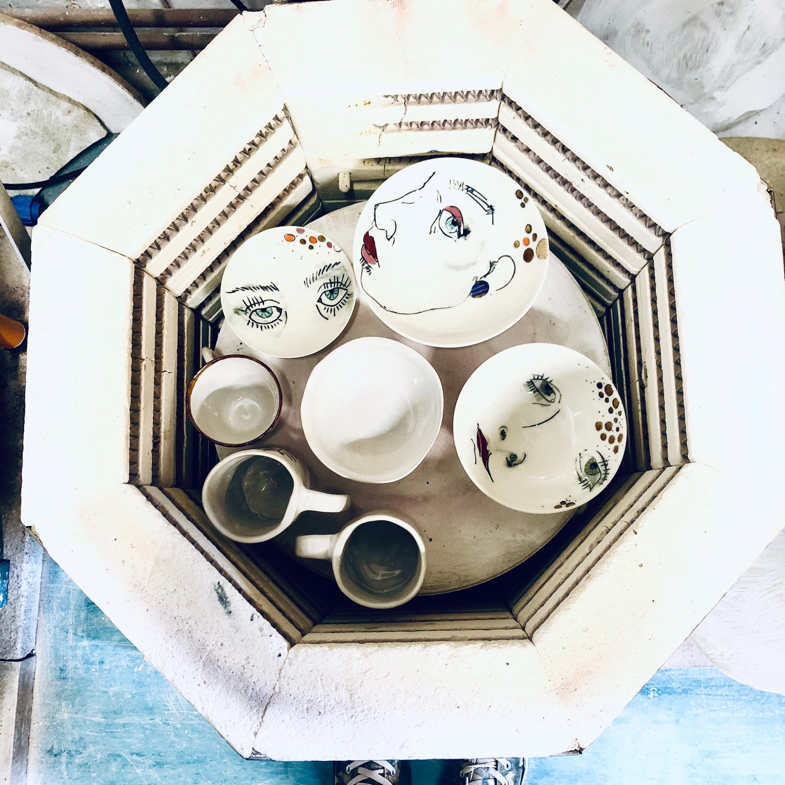 Top-down view of ceramic dishes and mugs with abstract face designs inside a hexagonal white structure.