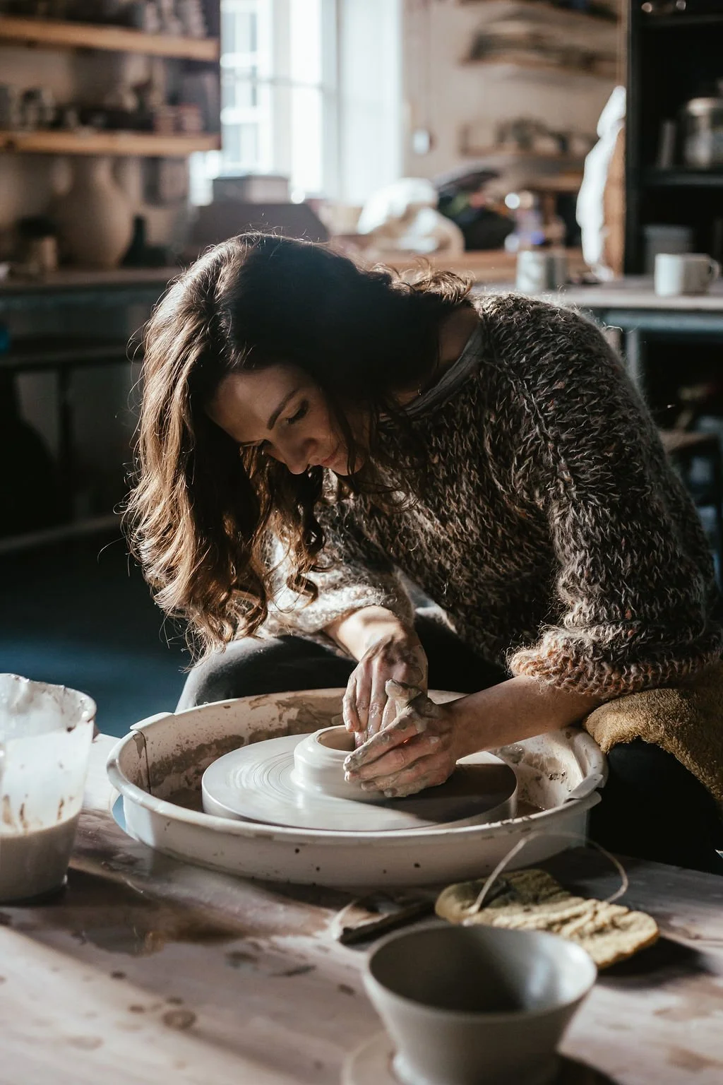 A woman with curly brown hair working on a pottery wheel in a studio, shaping a clay vessel.