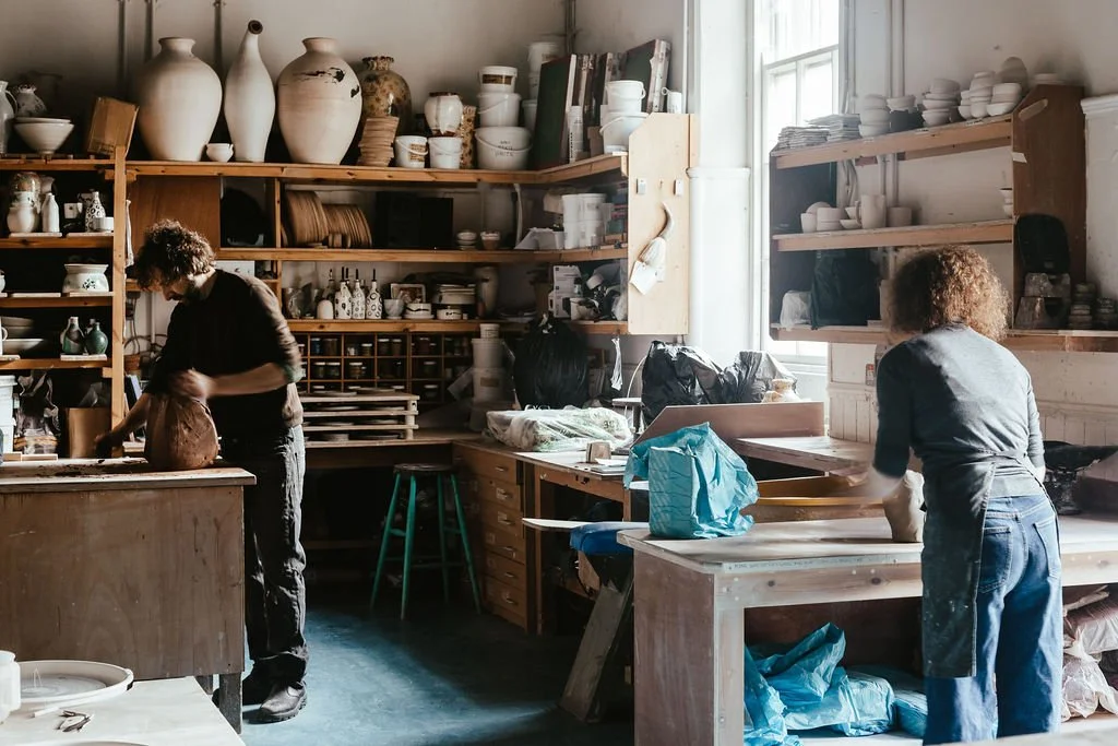 Two women working in a pottery studio with shelves of ceramic vases and bowls around them.