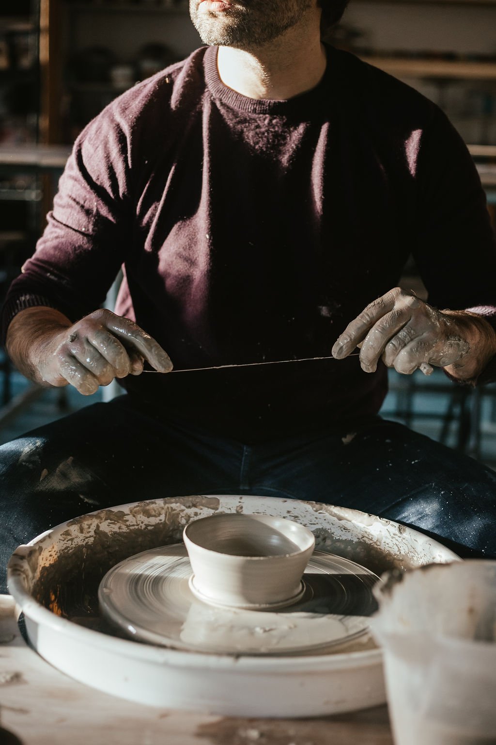 A man shaping a clay bowl on a pottery wheel in a studio. He is wearing a dark long-sleeved shirt and gloves, with pottery tools and supplies visible nearby.