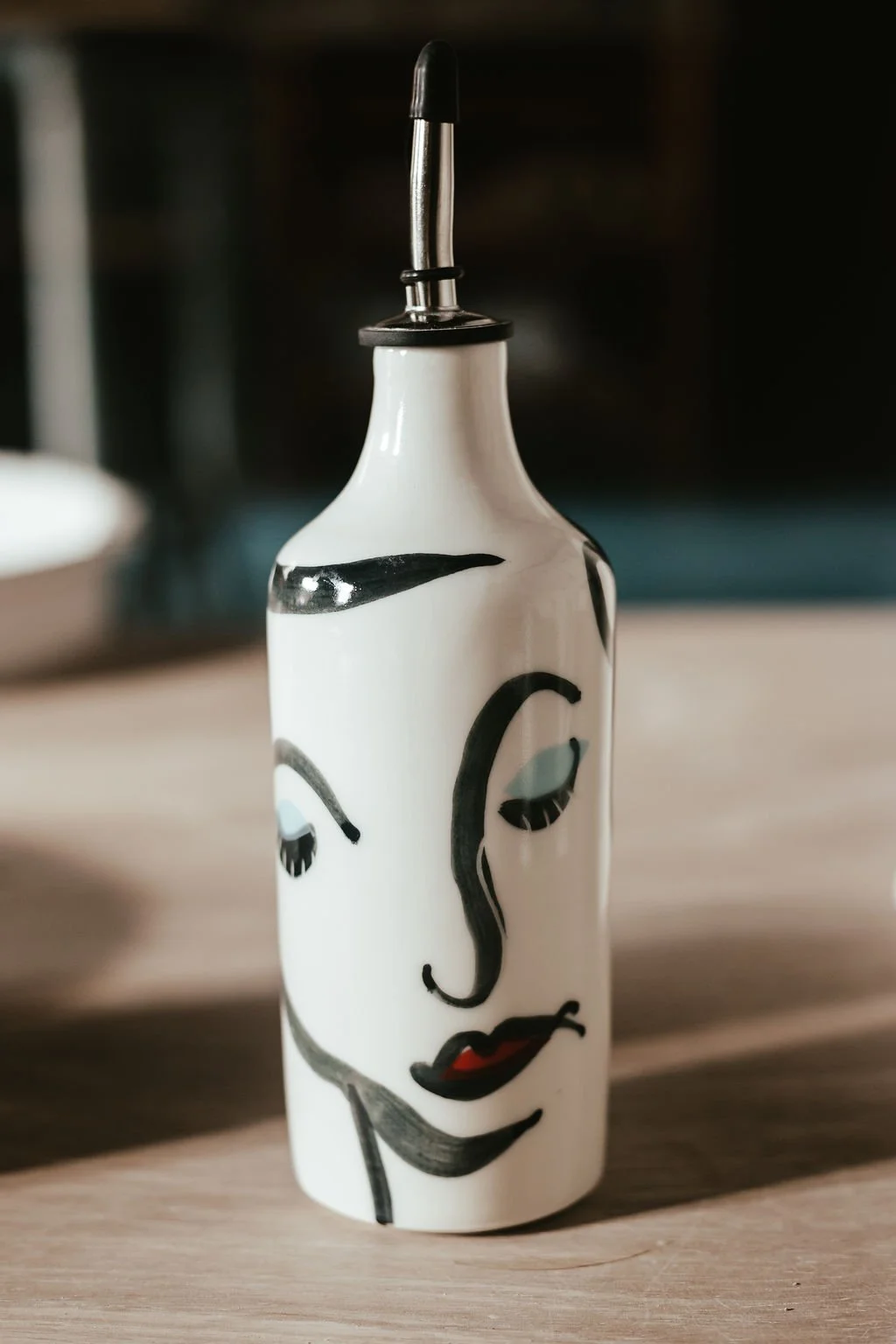 A ceramic squeeze bottle with black and red painted abstract face design, topped with a metal and rubber nozzle.