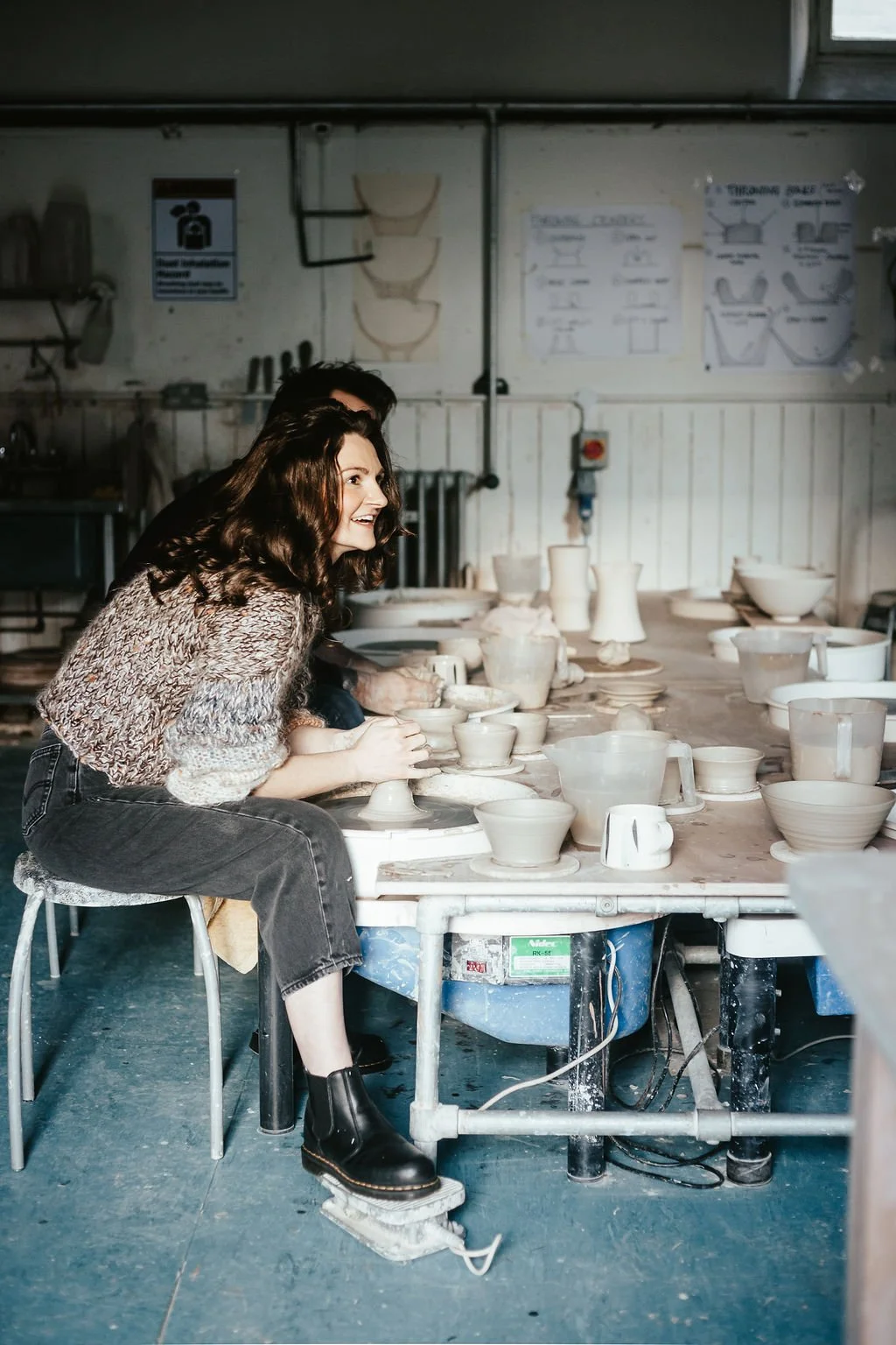 Woman with curly hair smiling and working on pottery at a pottery wheel in a ceramics studio, surrounded by finished and unfinished ceramic pieces.