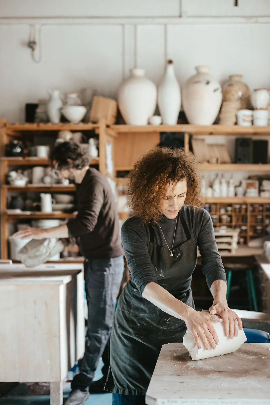 Two women working on pottery projects in a ceramics studio. One woman in the foreground is rolling out clay, while another in the background is working with clay on a table.
