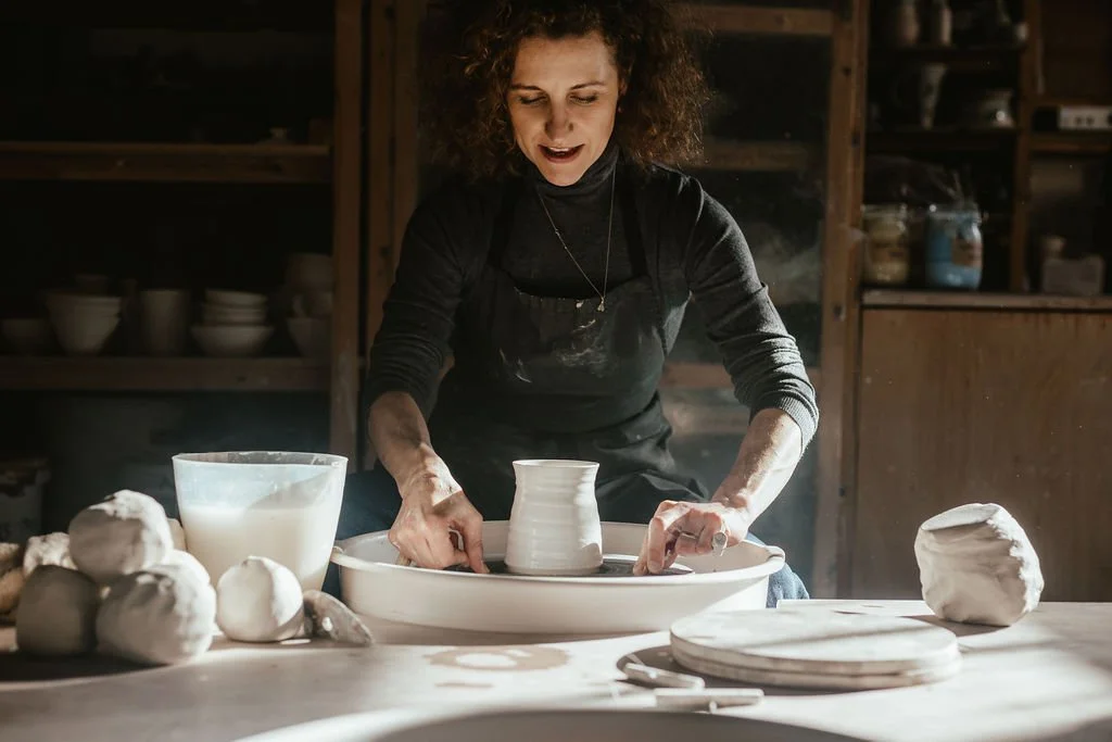 A woman making pottery on a spinning wheel in a ceramics studio, surrounded by clay and pottery tools.