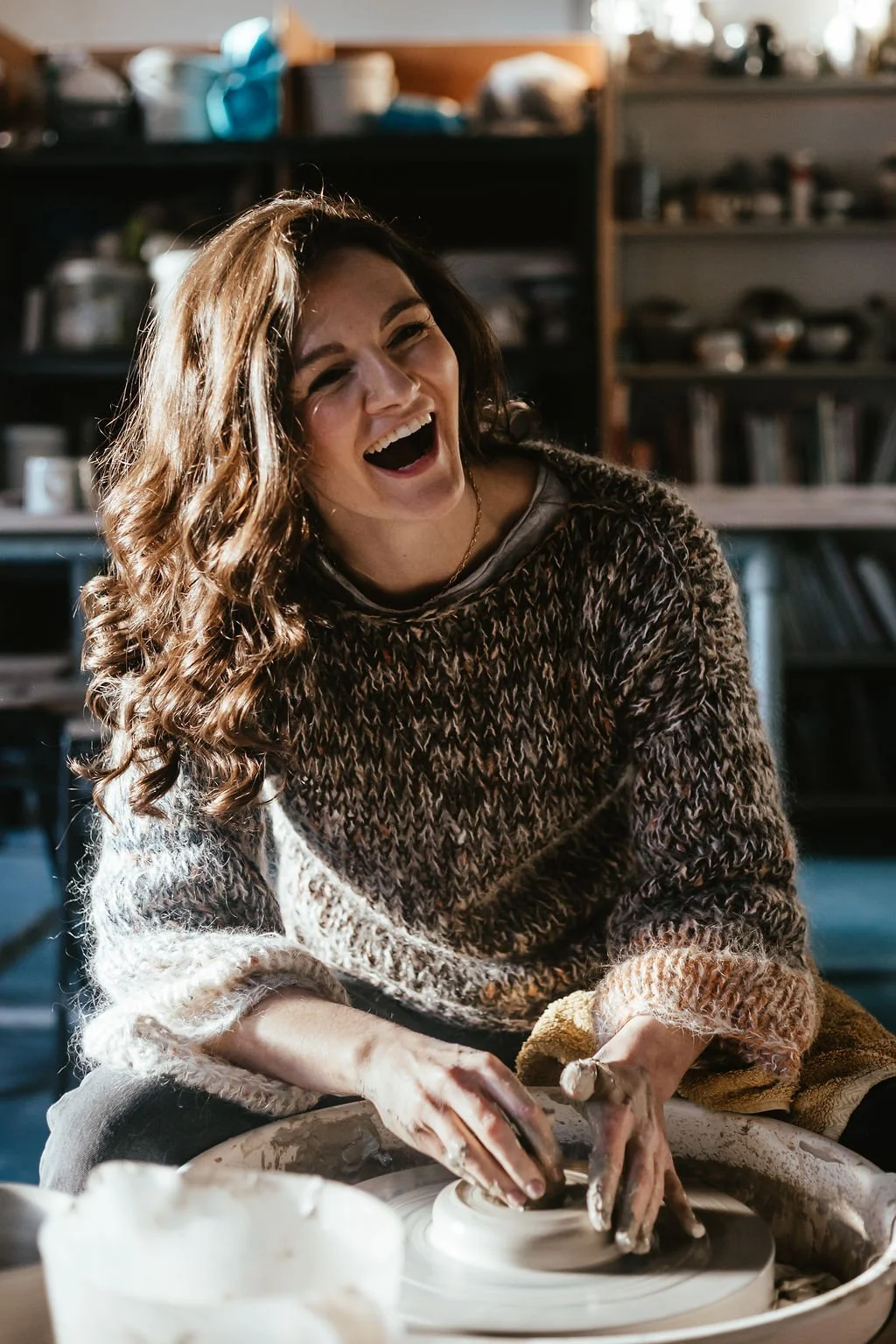 A young woman with curly hair is laughing while working with clay on a pottery wheel in a cozy, well-lit studio.