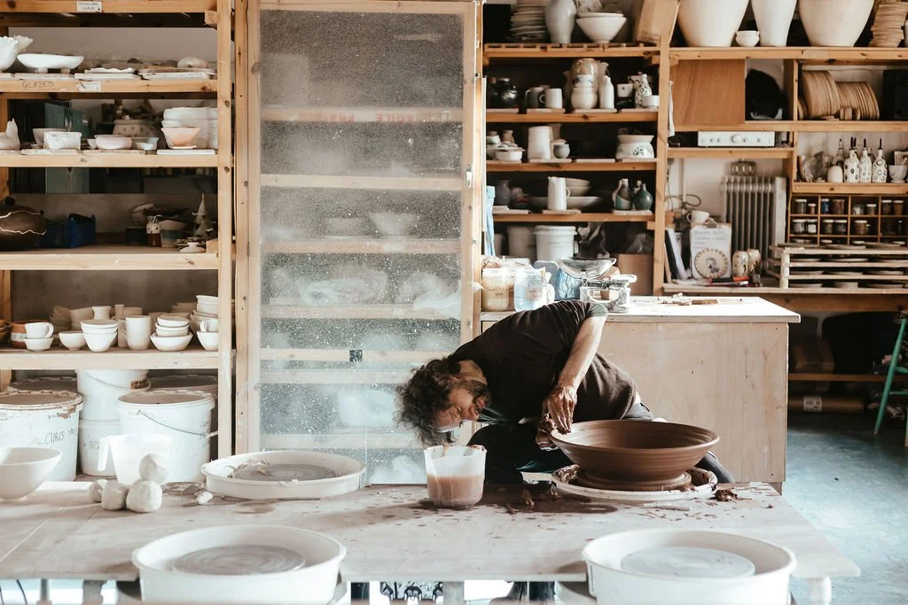 A ceramic artist working on a large bowl in a pottery studio filled with shelves of pottery pieces and tools.