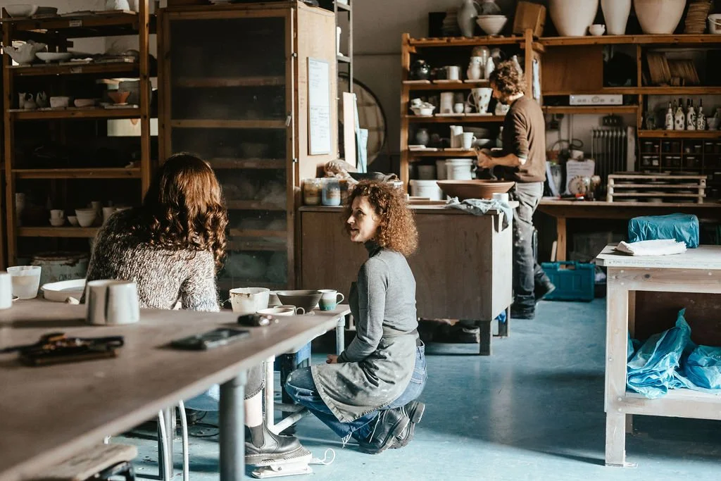 Two women are sitting and talking in a pottery studio with shelves of pottery and clay tools behind them, while a person in the background is working on pottery.