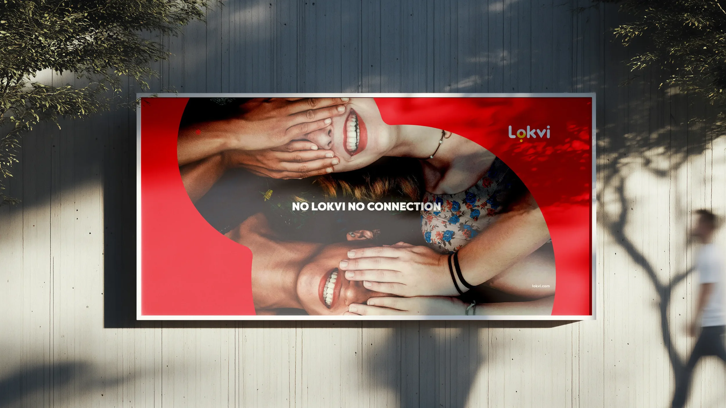A billboard featuring four smiling women with their faces covered by hands, set against a red background. The billboard has the text "NO LOKVI NO CONNECTION" and the logo "Lokvi" in the top right corner. The billboard is mounted on a white wall, with a person walking nearby and a tree casting shadows.