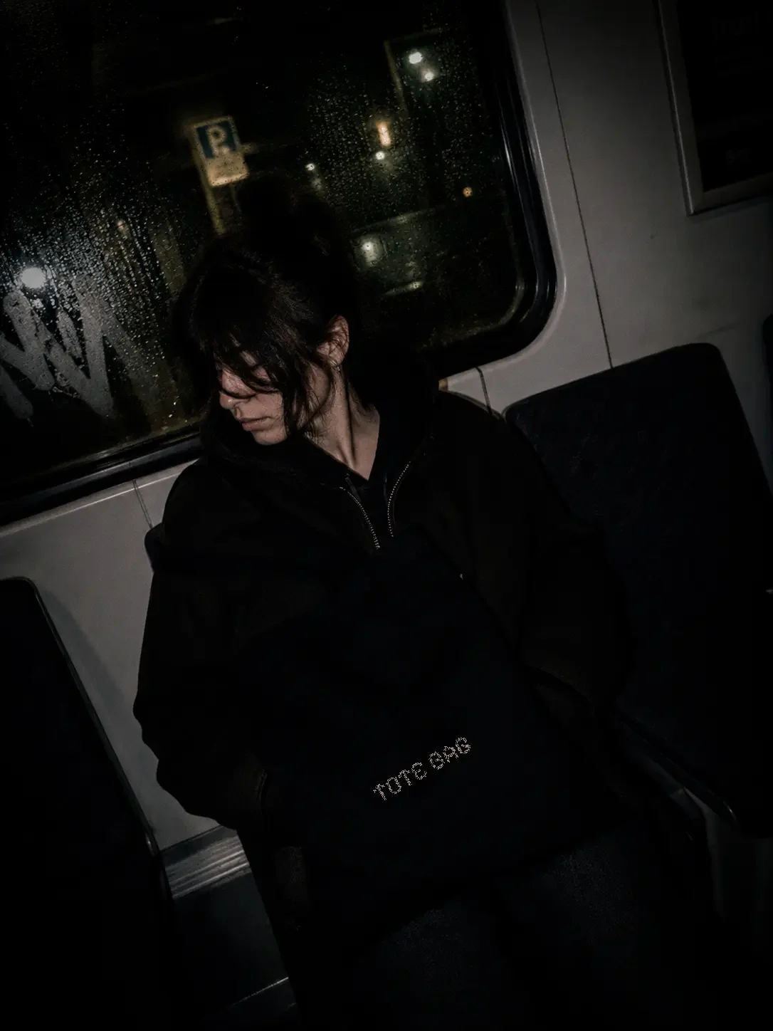 A woman with dark hair and glasses sitting alone on a bus at night, looking down, with rain on the window behind her.